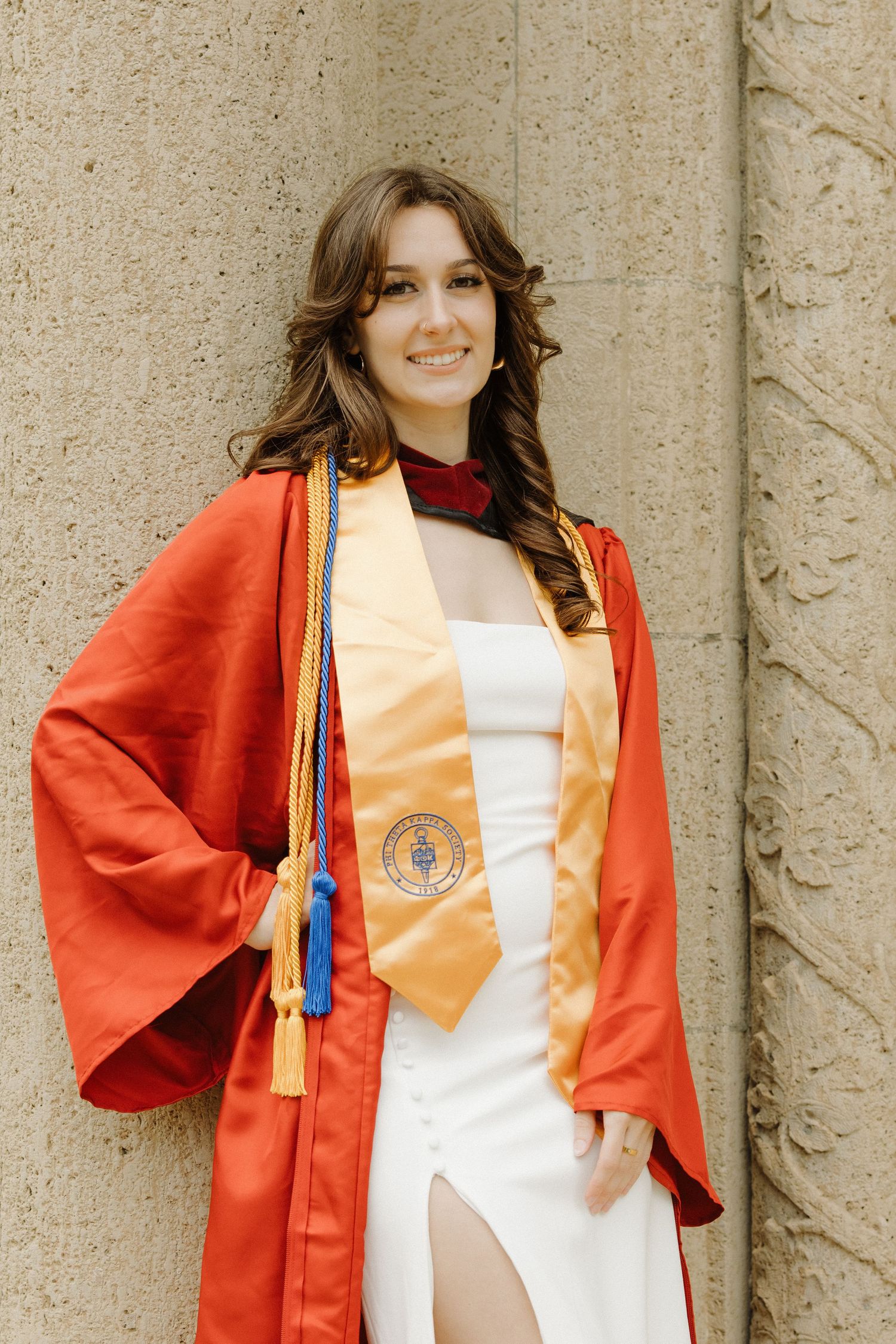 Graduate poses in orange academic regalia and white dress against stone column.