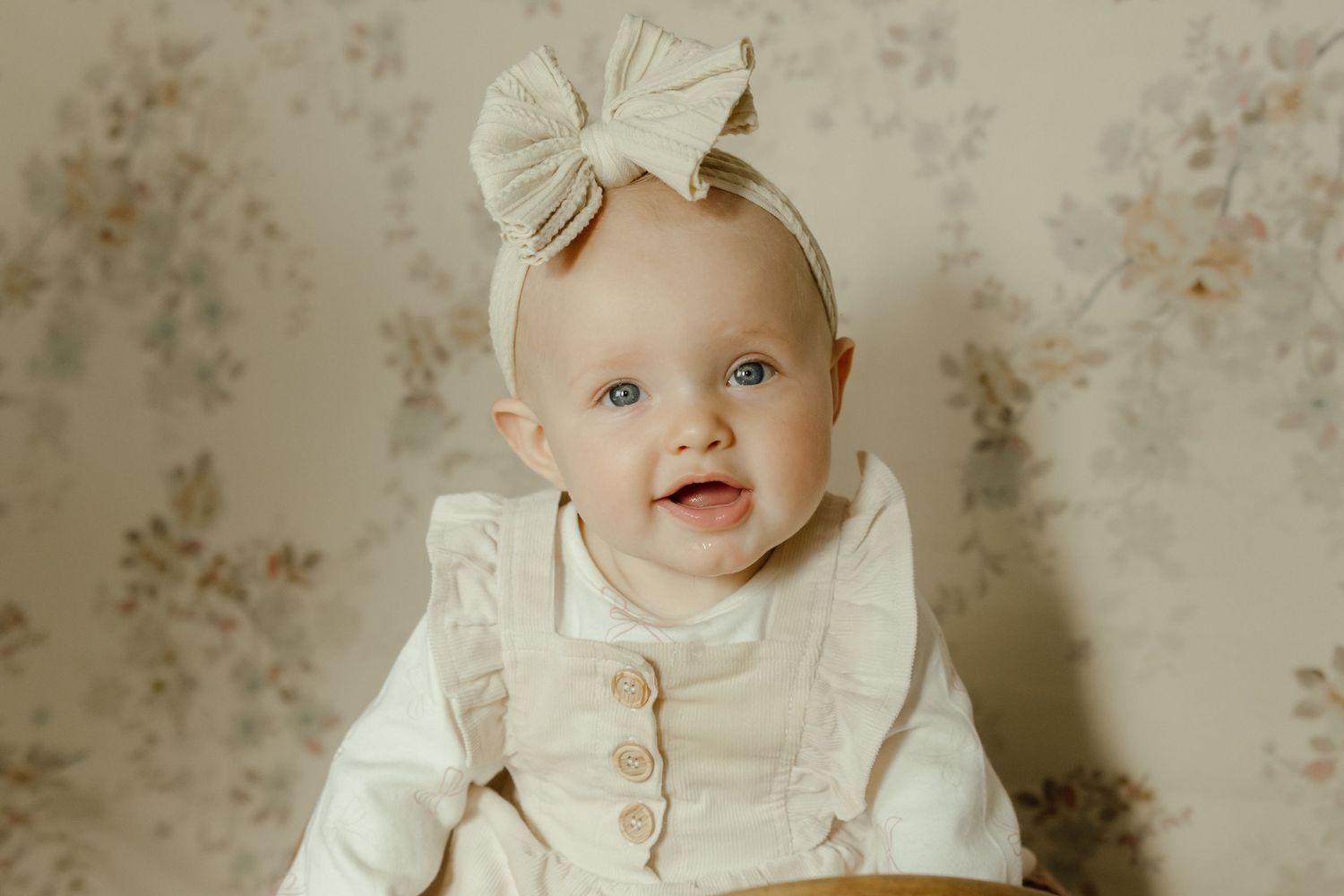 Sweet baby in vintage-style cream dress and bow poses in front of floral wallpaper in studio setting.