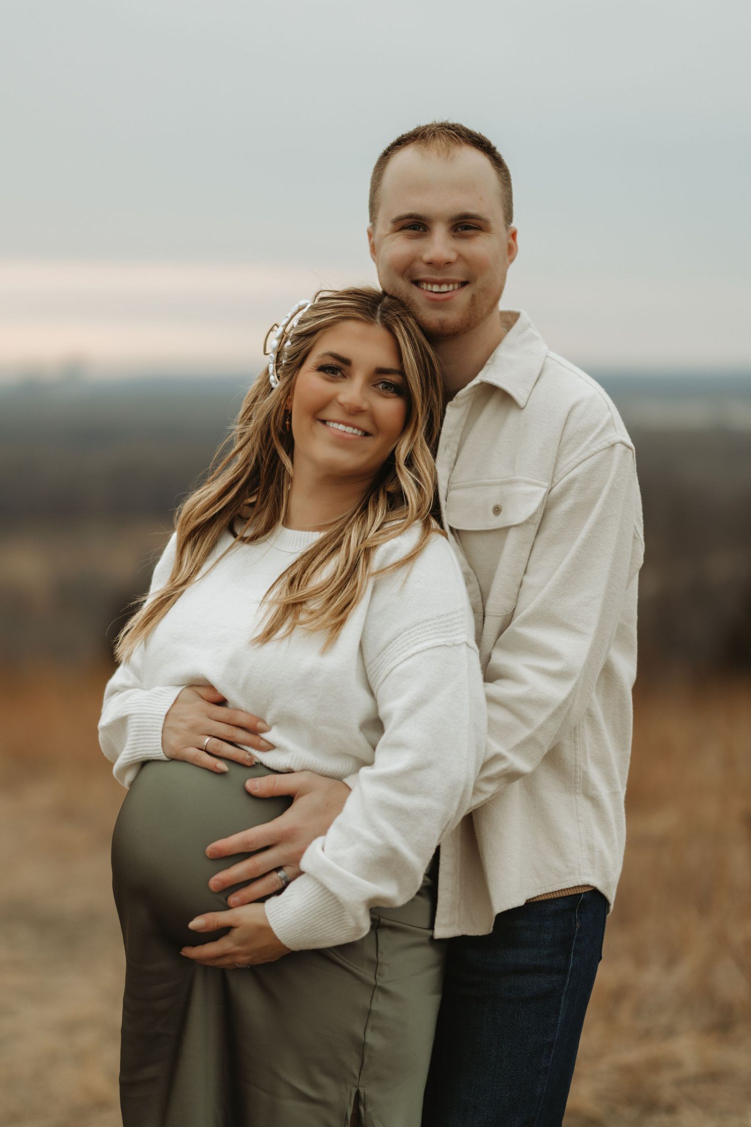 A couple embraces in a field during a maternity photo session wearing white and olive green outfits at sunset.