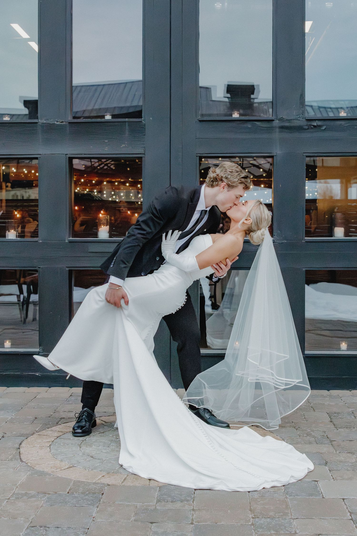 Groom dips bride in elegant white gown and veil for a romantic kiss outside a venue with string lights.