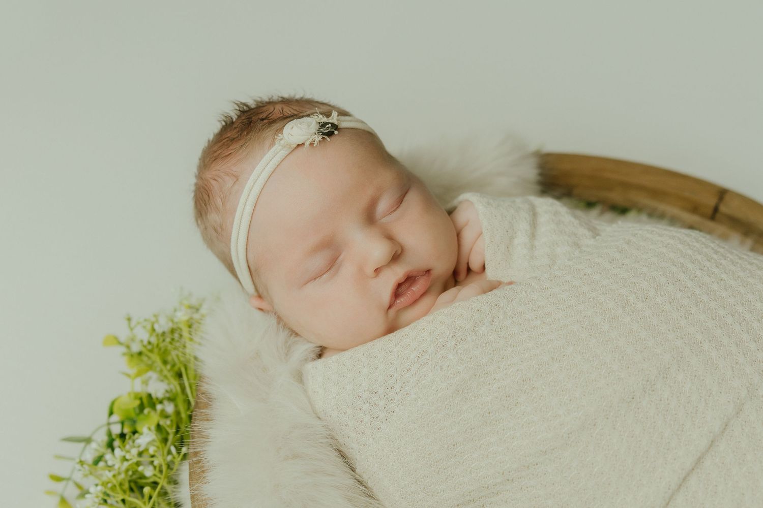 Peaceful newborn baby girl sleeping swaddled in cream wrap with floral headband in a wooden bowl.