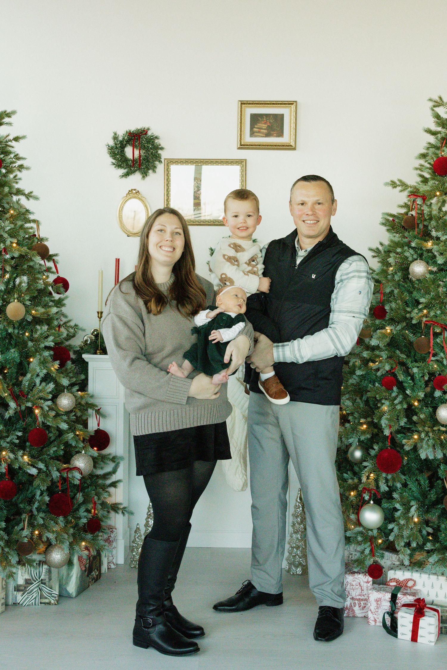 Family of three posing by decorated Christmas trees with gifts, parents holding toddler in festive holiday home setting.