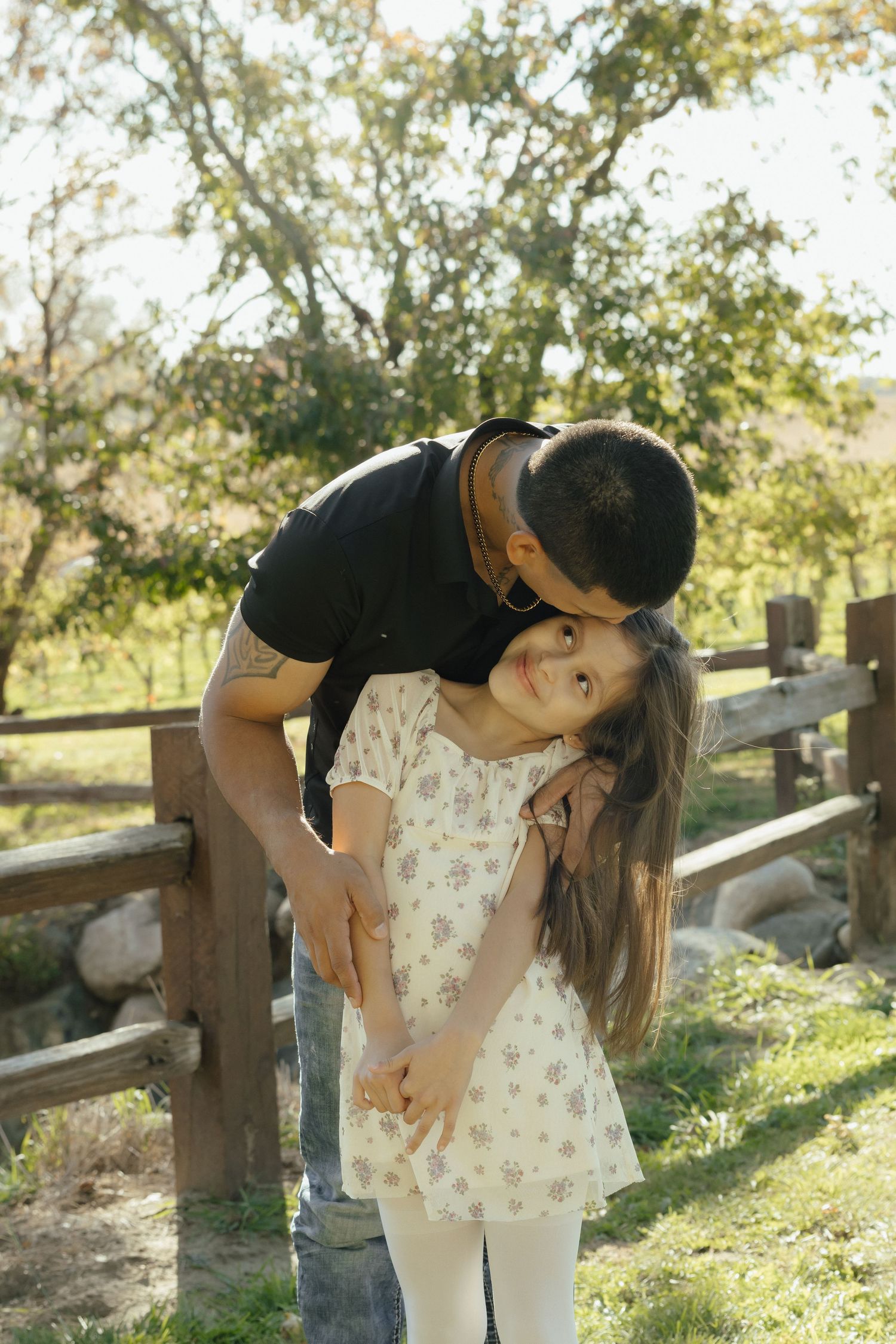 A father lovingly kisses his young daughter on the head outdoors near a wooden fence.