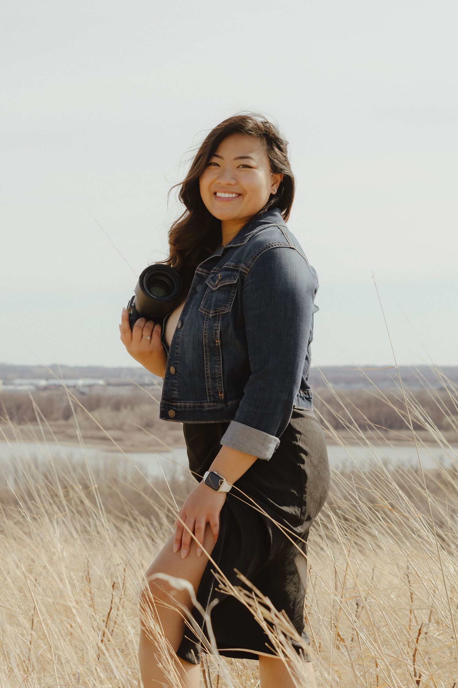 A person in a denim jacket poses playfully in a field of tall golden grass on a bright sunny day.