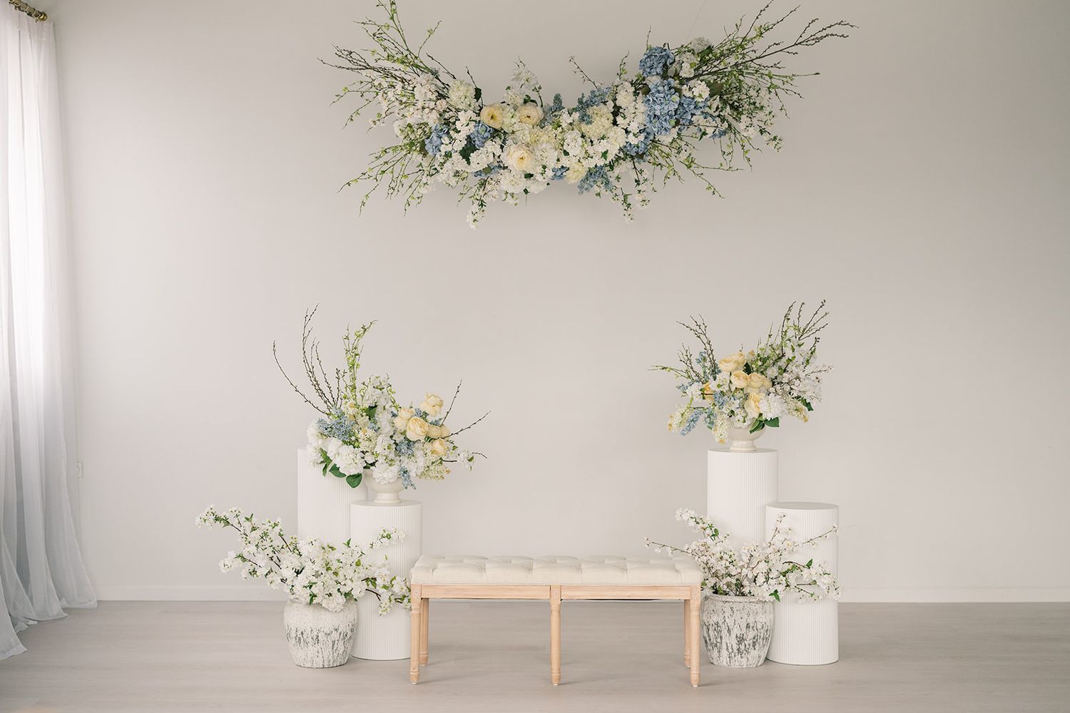 Elegant white room with floral arrangements of blue and white flowers, a wooden bench, and a wall-mounted bouquet.