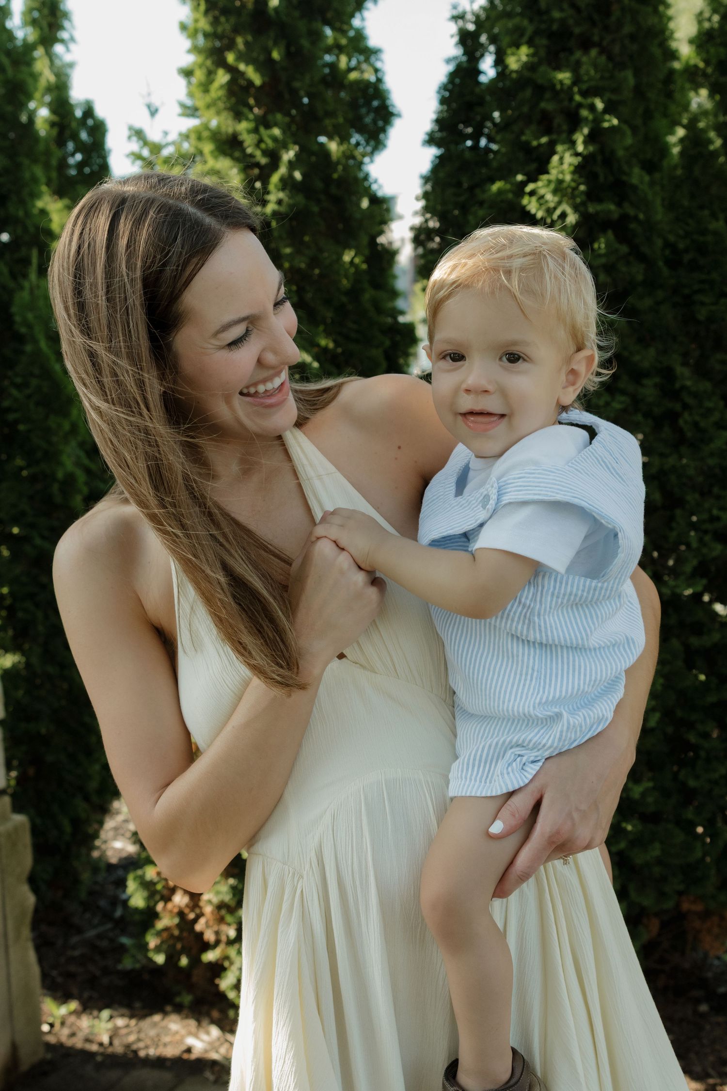 Smiling mother in white dress holding toddler boy in light blue outfit outdoors near green trees.