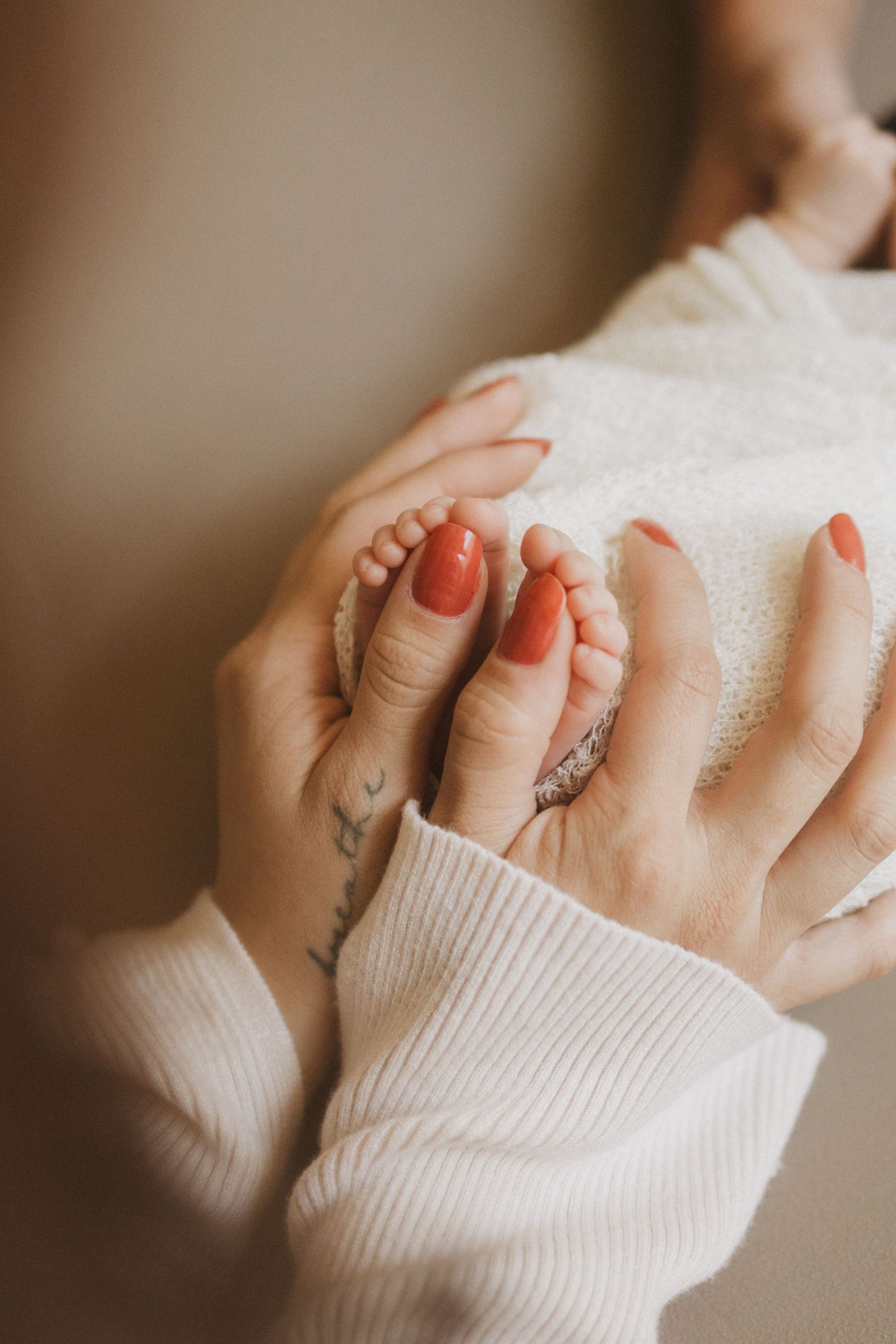 A mother with red nails and a tattoo gently holds her newborn baby's tiny feet in her hands.