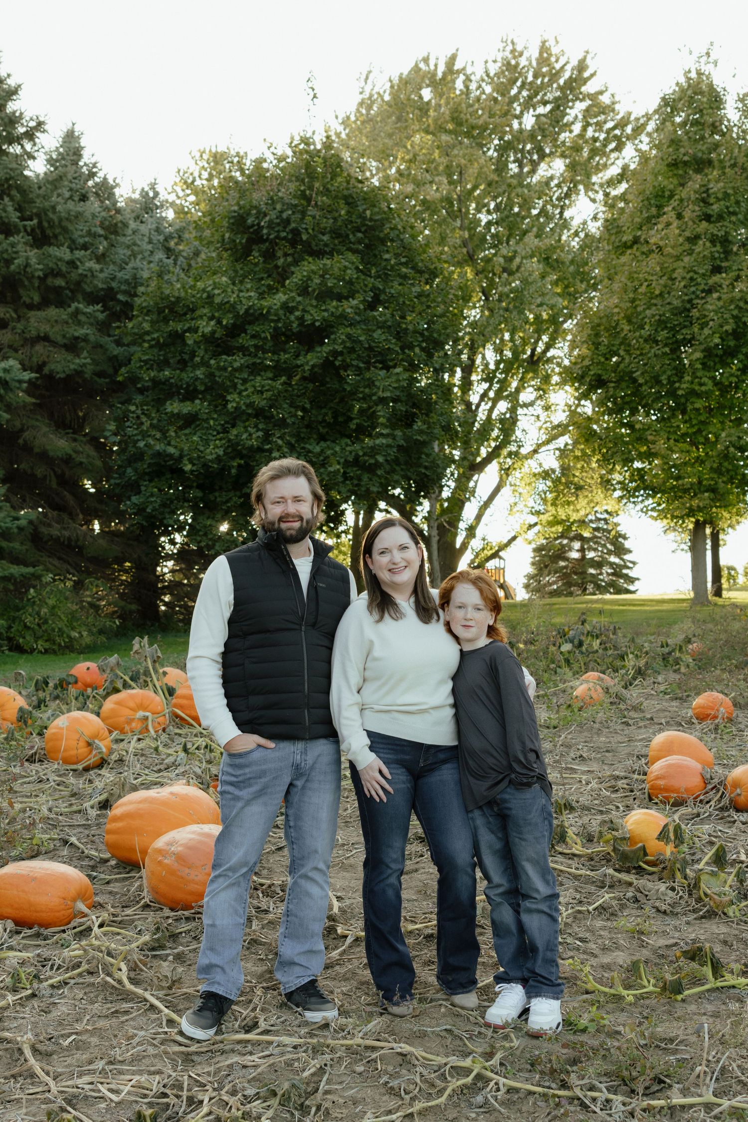 Family of three smiling in a pumpkin patch surrounded by orange pumpkins and green trees on a fall day.
