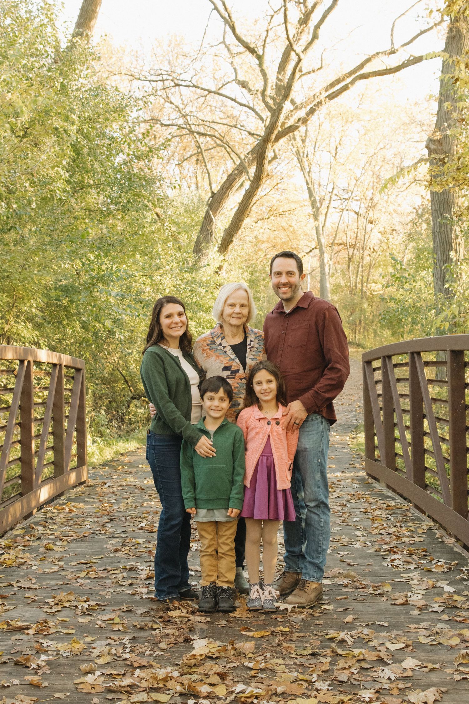 Family of five posing on a wooden bridge surrounded by autumn foliage in a park setting.