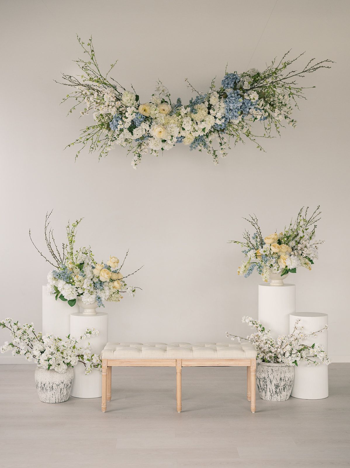 Elegant wedding backdrop with blue and white floral arrangements on white pedestals and a tufted bench against a neutral wall.