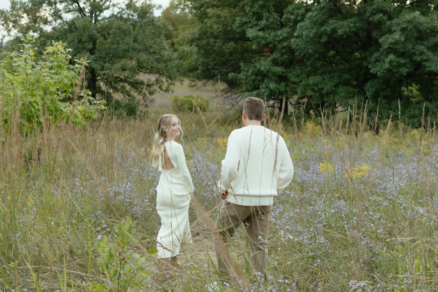 Two people walk through a wildflower meadow with tall grass and purple flowers on a summer day.