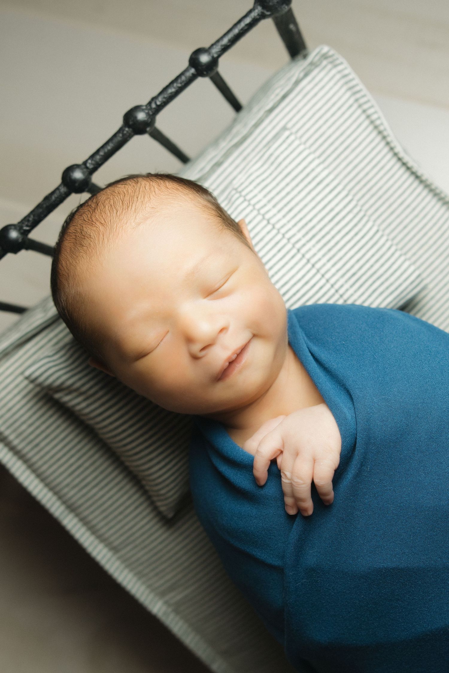 Peaceful newborn baby swaddled in blue wrap sleeping on miniature striped bed during professional newborn photo session.