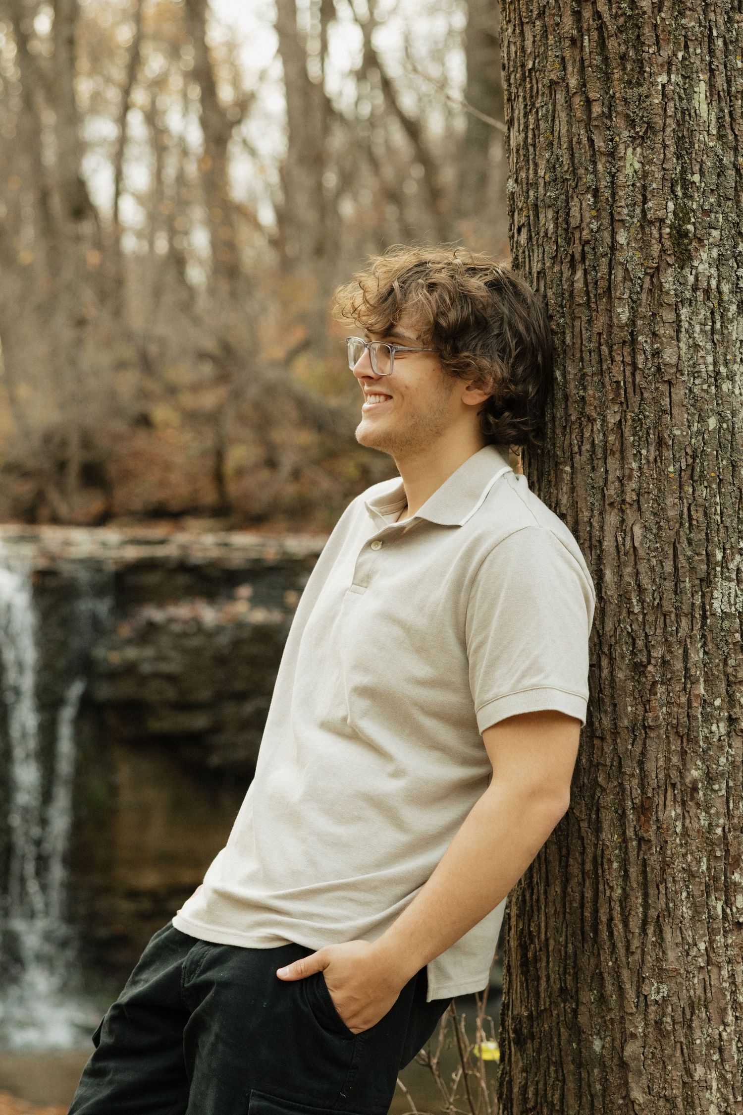 A person in a white polo shirt leans against a tree trunk with a waterfall visible in the background.