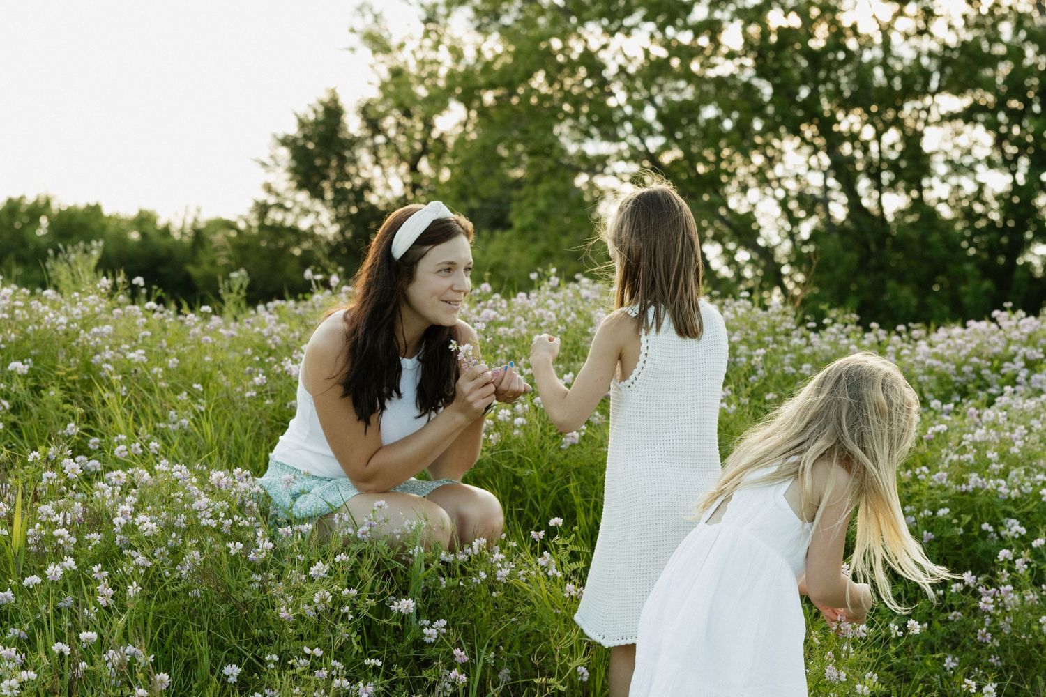 Woman crouching in wildflower meadow with two young girls in white dresses on a sunny day.