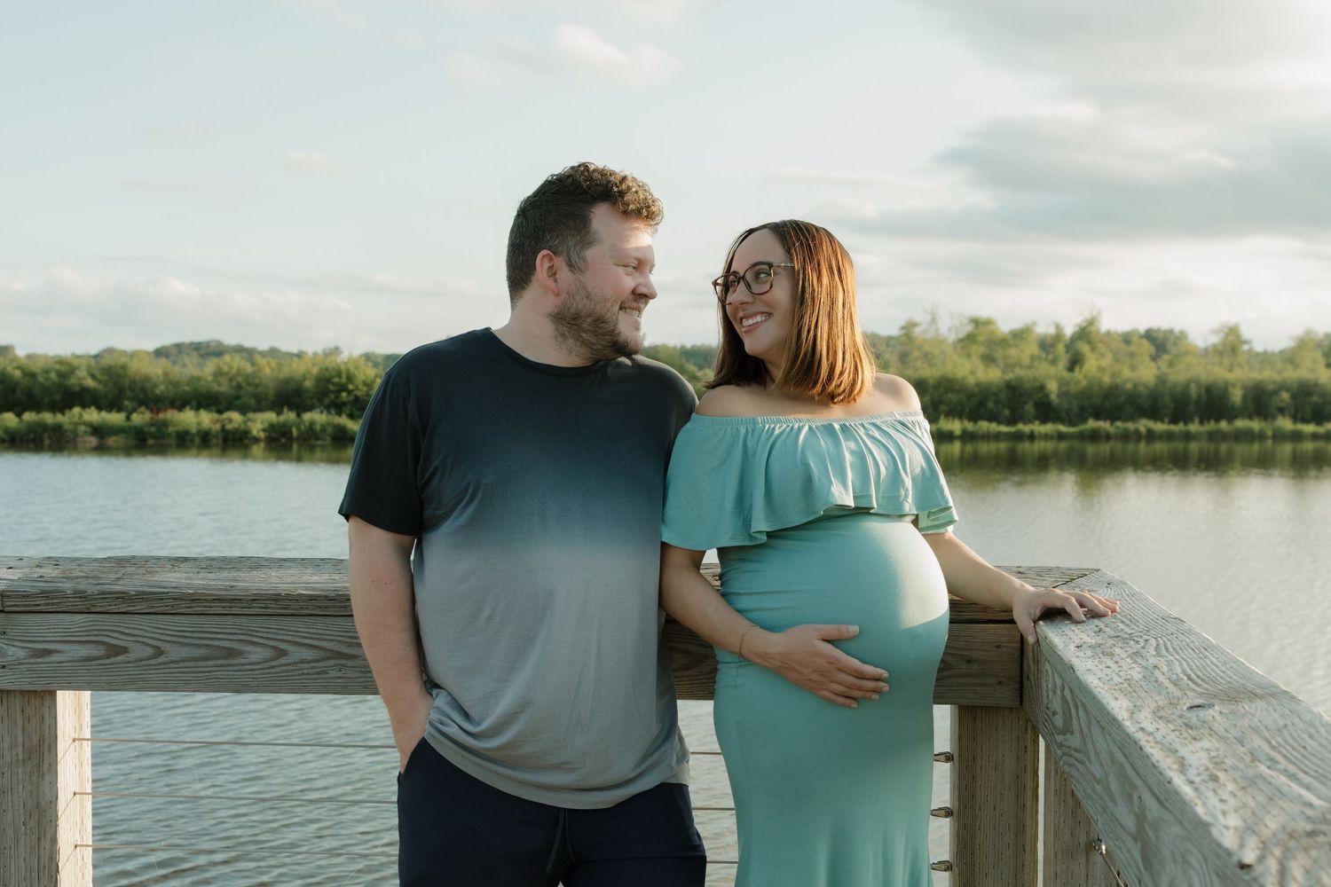 Couple smiling on wooden dock by lake, pregnant woman in teal off-shoulder dress cradling baby bump.
