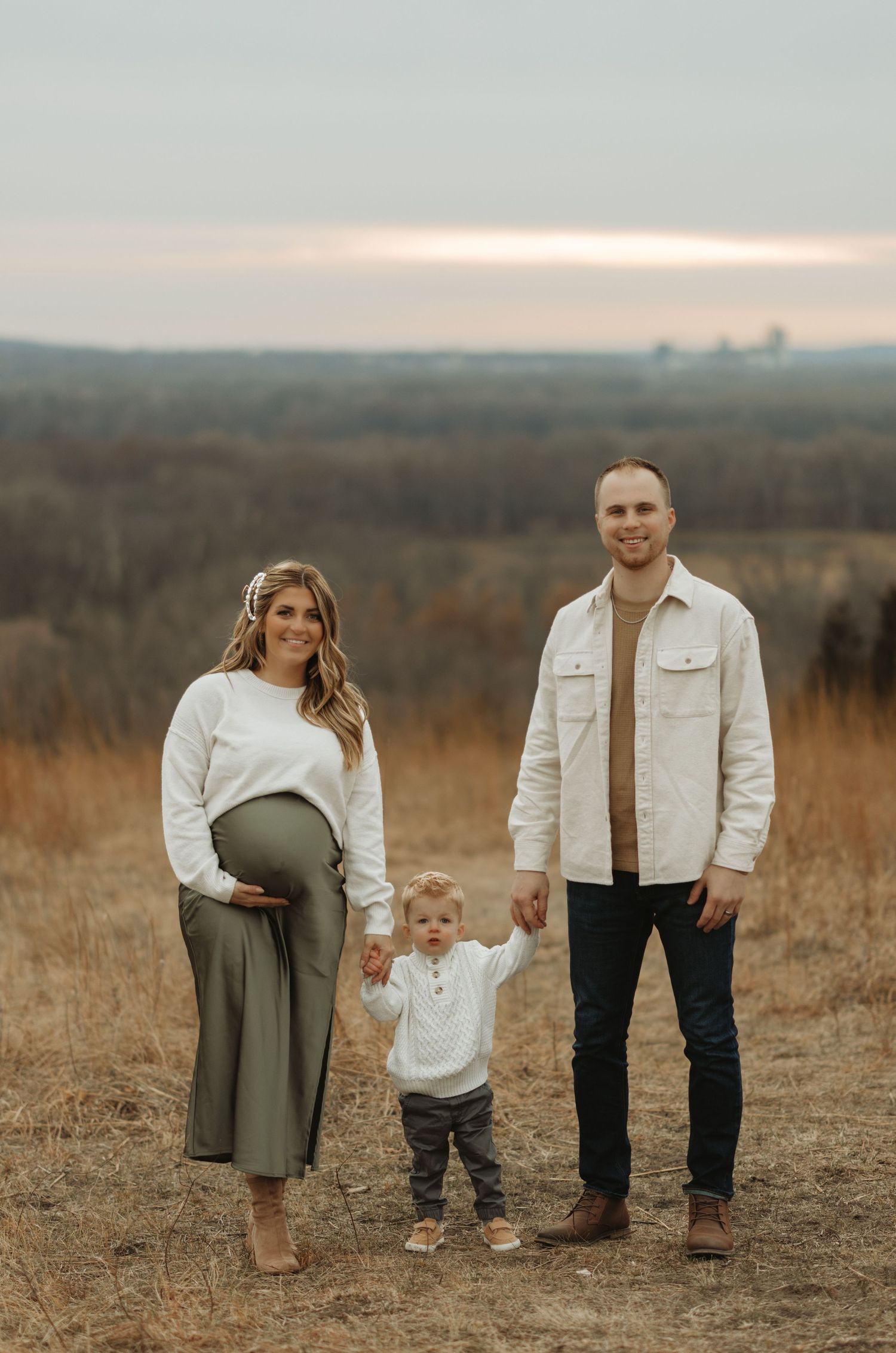 Family stands together in field during sunset with rolling hills in background.