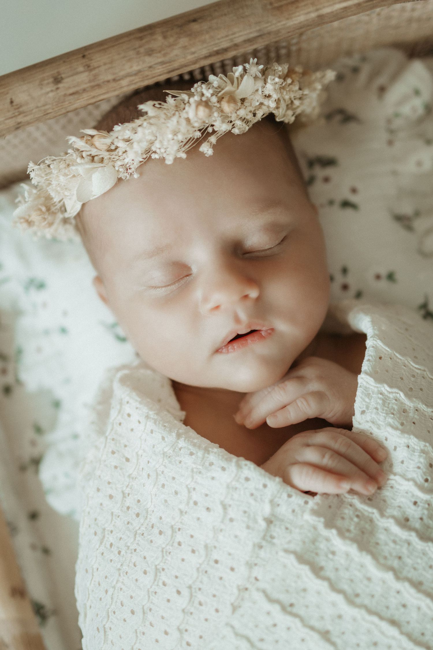 Sleeping newborn baby girl wearing a delicate floral crown, wrapped in a white lace blanket in a wooden bassinet.
