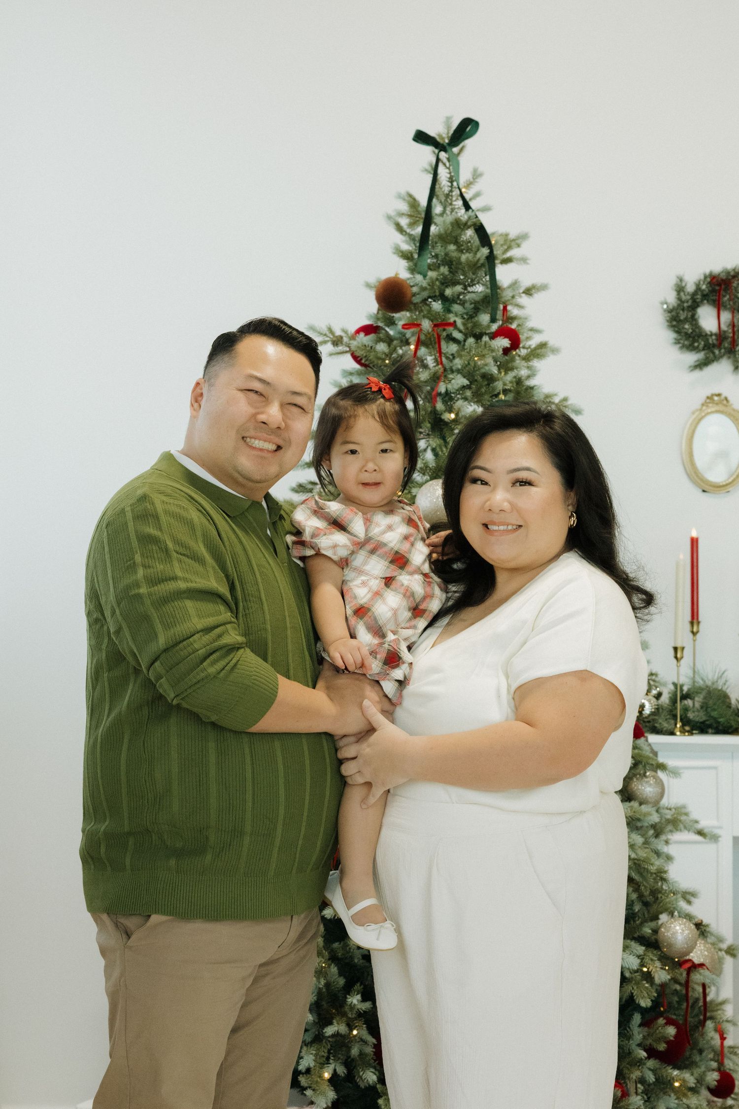 Family of three posing in front of decorated Christmas tree, parents holding young daughter in festive holiday attire.
