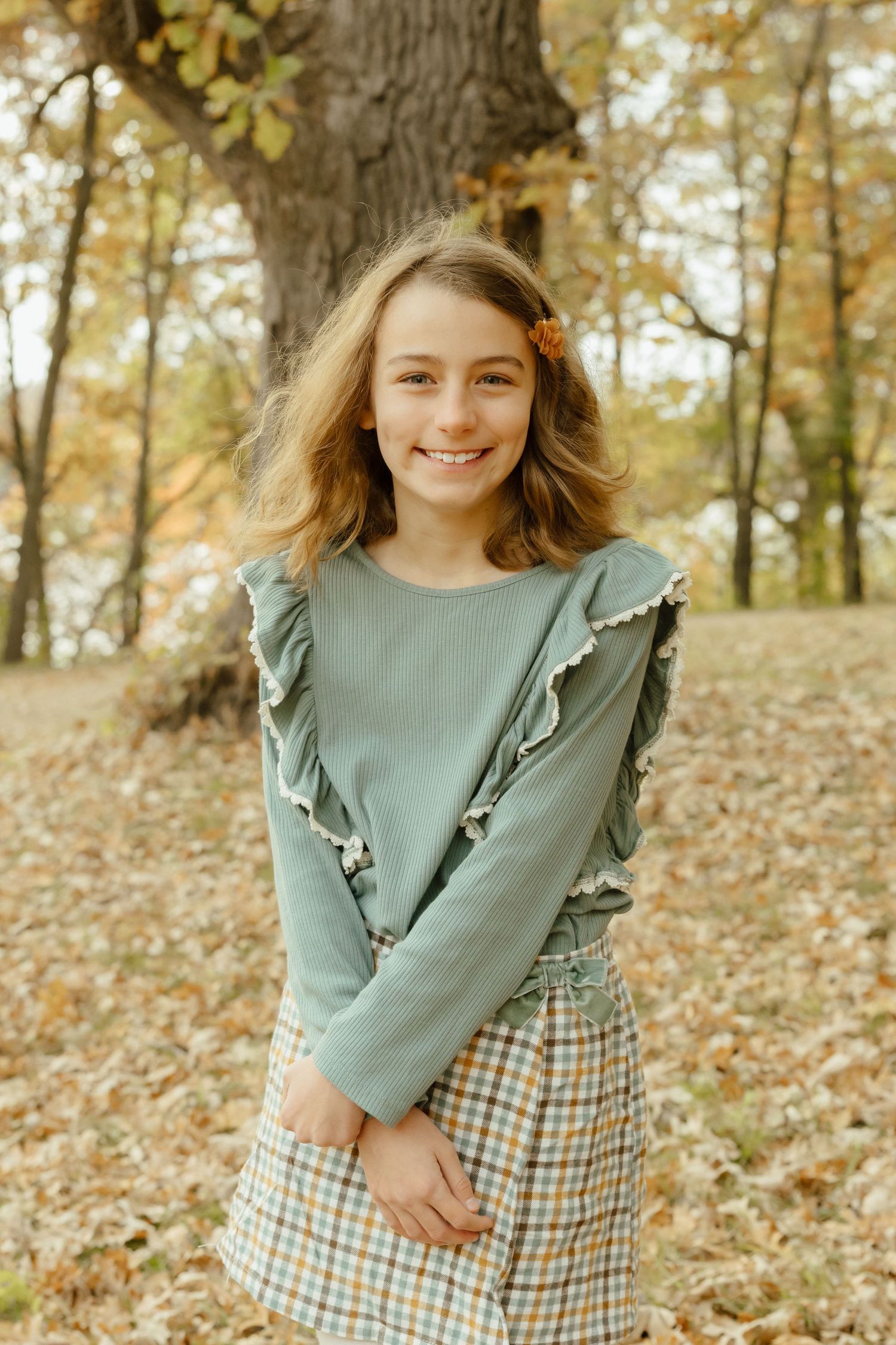 Smiling young girl in teal ruffled top and plaid skirt standing among autumn leaves in a forest park.