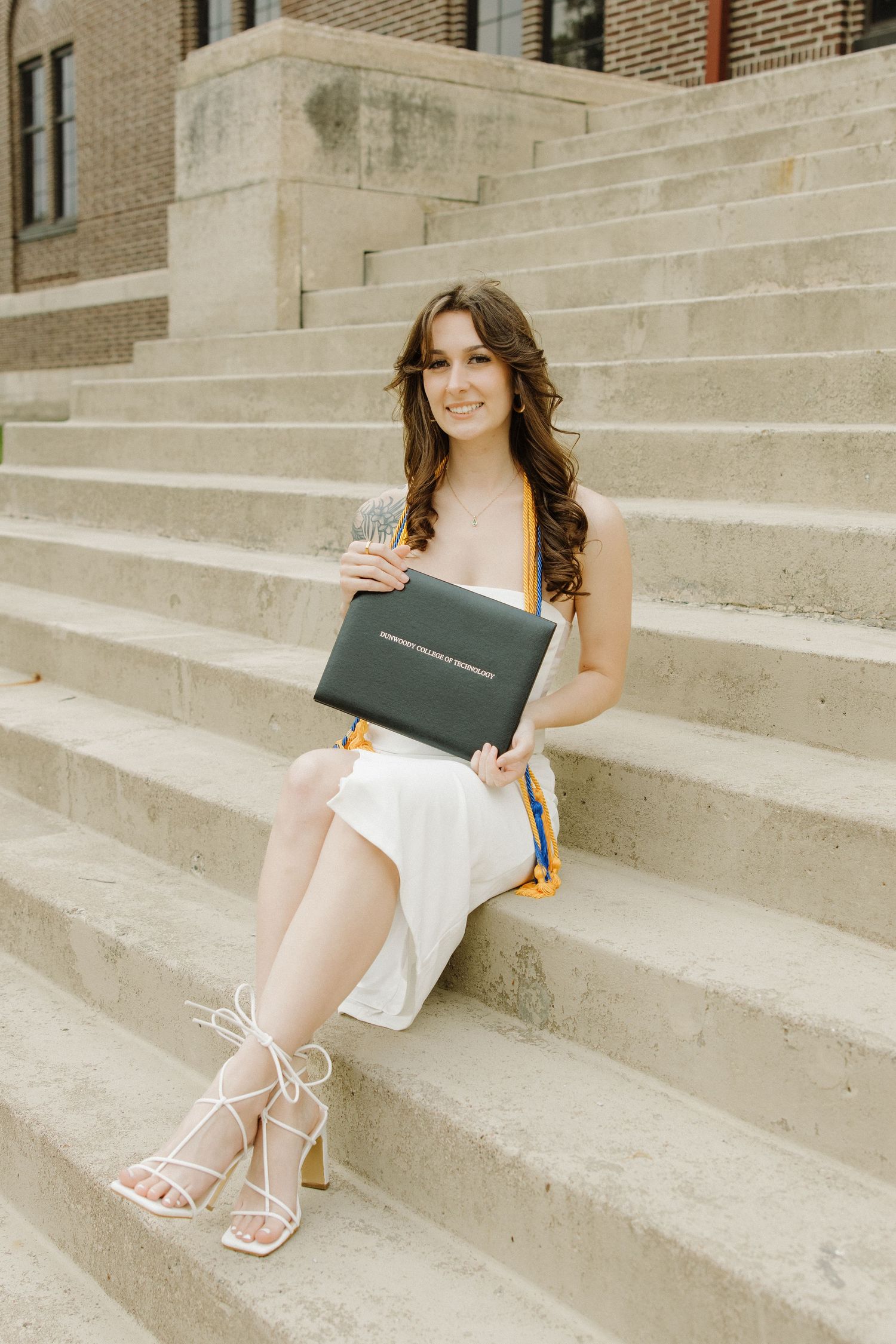 Graduate in white dress sits on concrete steps holding diploma while wearing strappy heels.