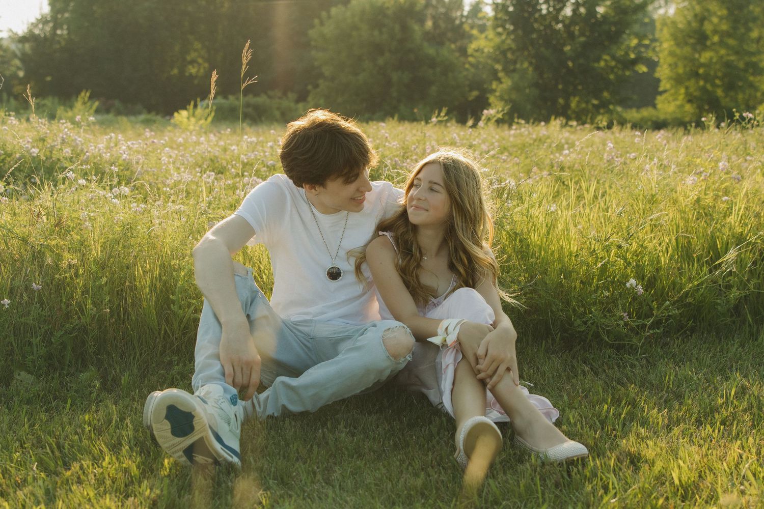 Couple in white outfits sits together in a sunlit meadow with golden wildflowers and grass.