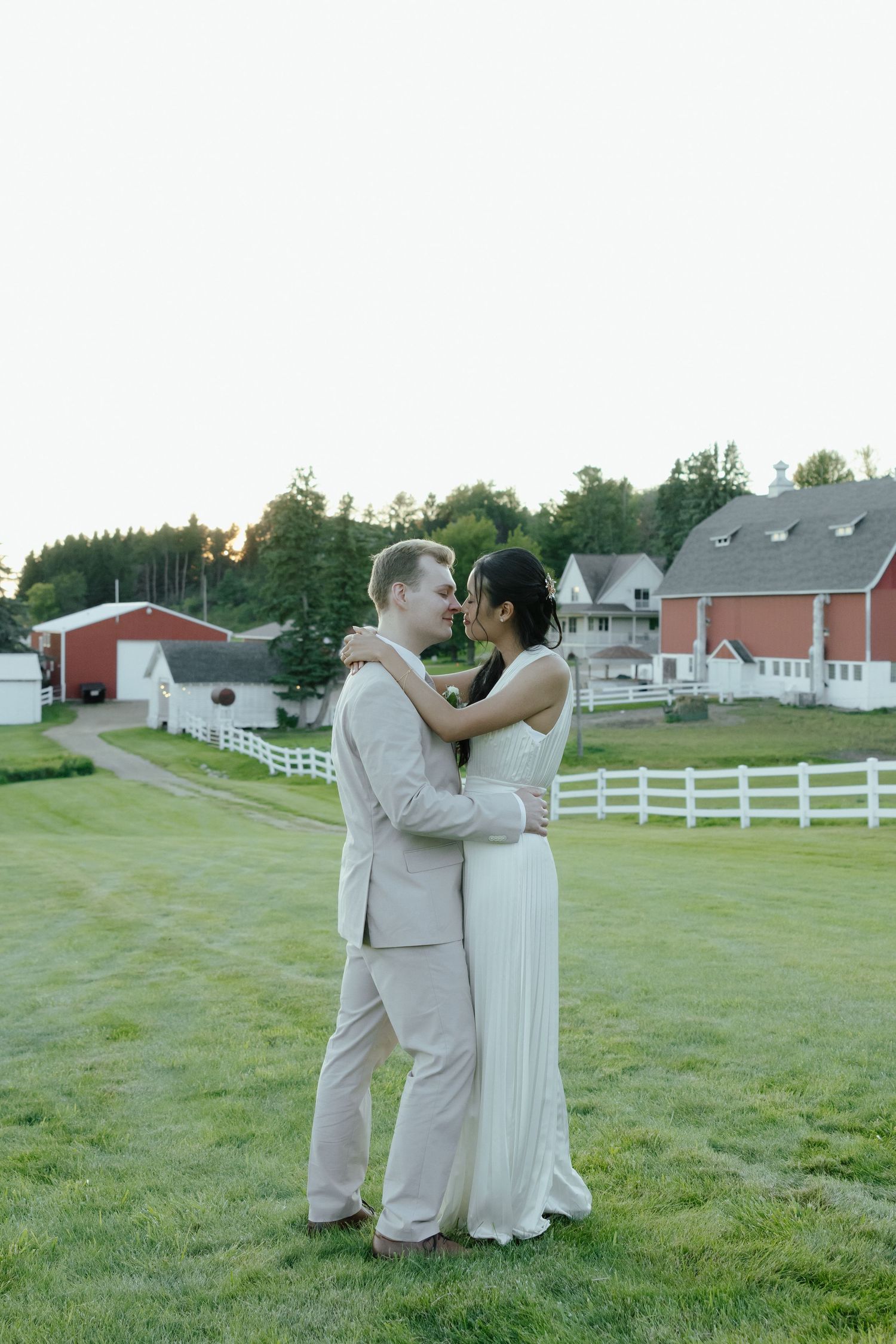 A couple shares a romantic moment on a grassy lawn with white fences and red barns in the background at sunset.