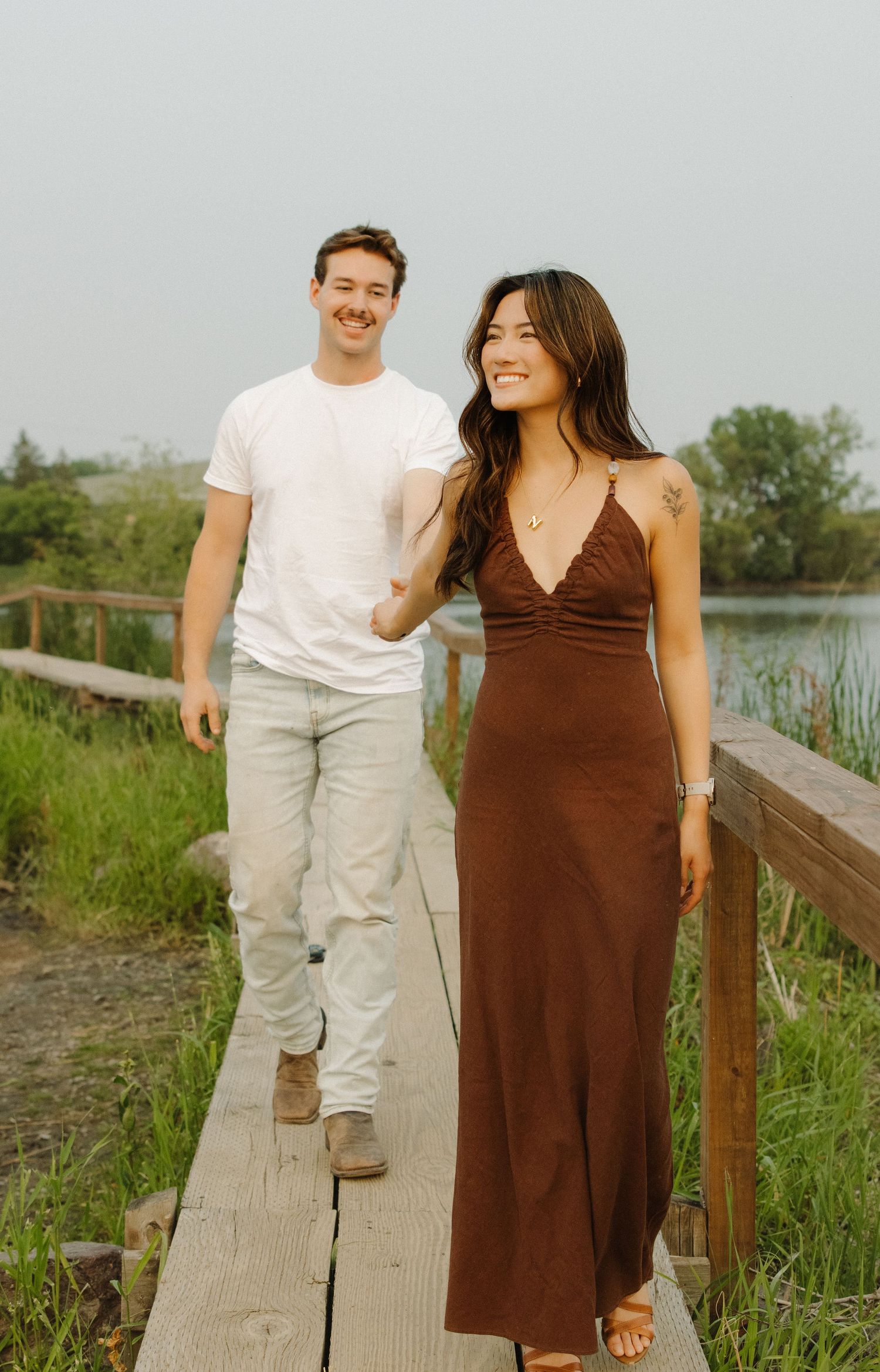 A couple walks hand in hand along a wooden boardwalk surrounded by green grass and marsh vegetation.