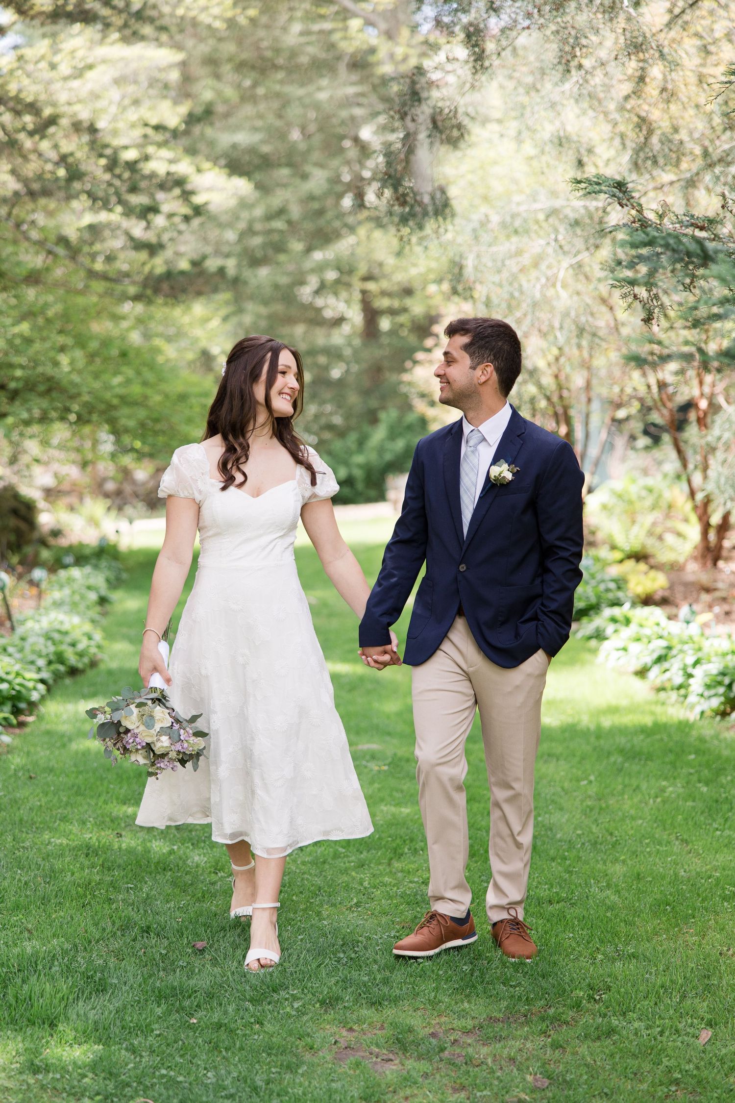A couple walks together on lush grass, with the bride in a midi dress and groom in navy blazer.