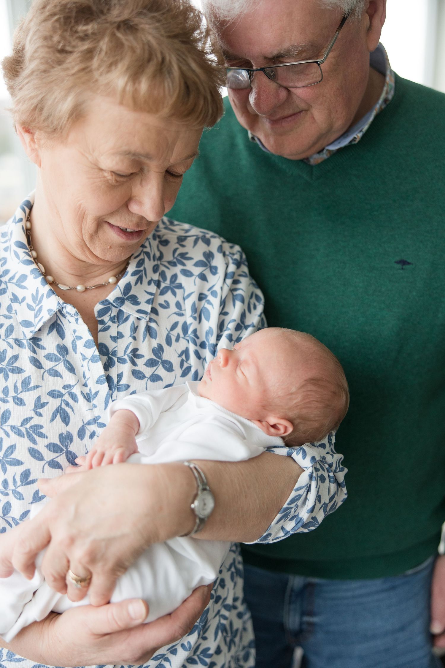 A grandparent couple in casual attire lovingly holds and gazes at a sleeping newborn baby dressed in white.