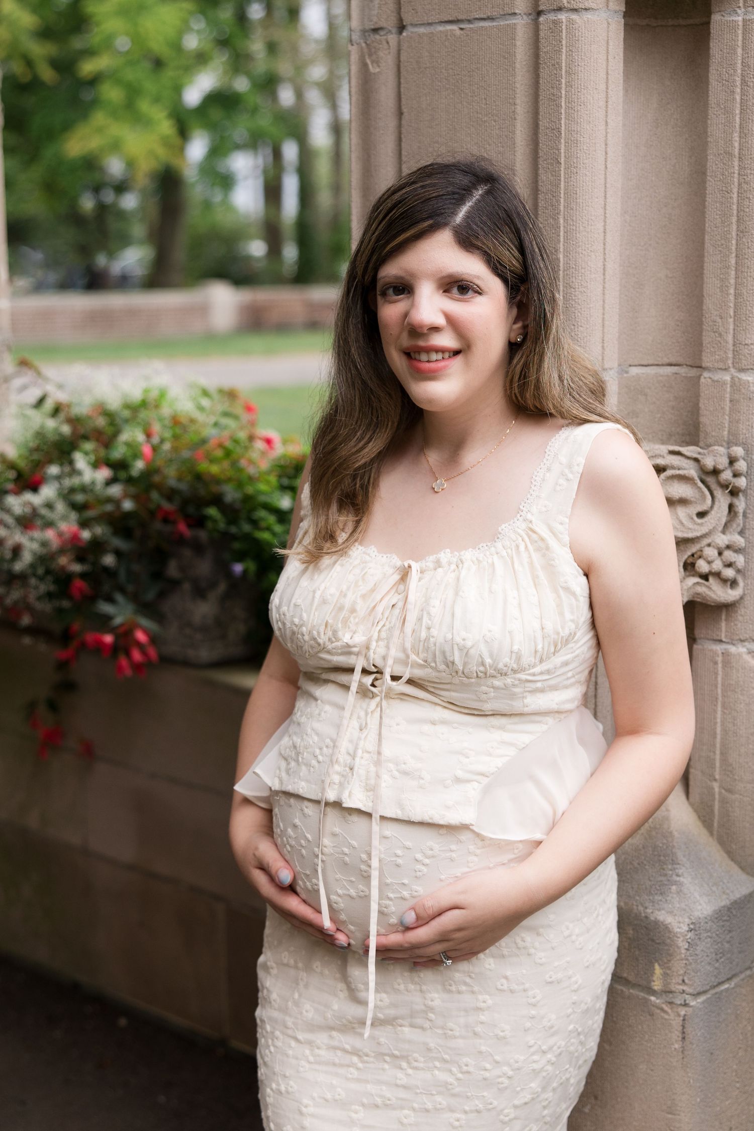 Expectant mother in a white vintage-style dress poses by stone architecture and flowering bushes.
