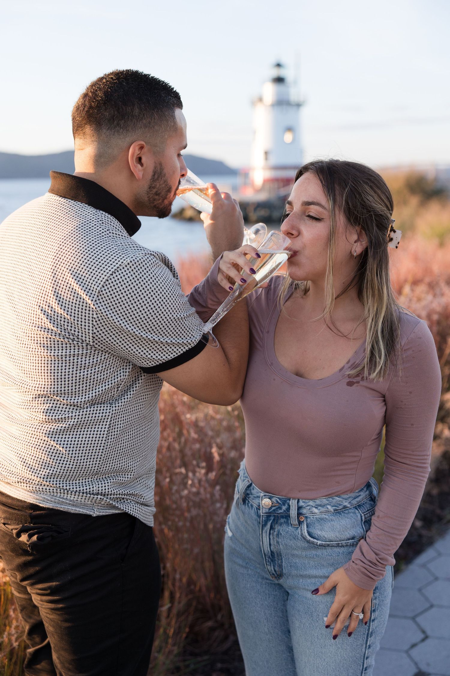 A couple drinks champagne together near a lighthouse at golden hour.