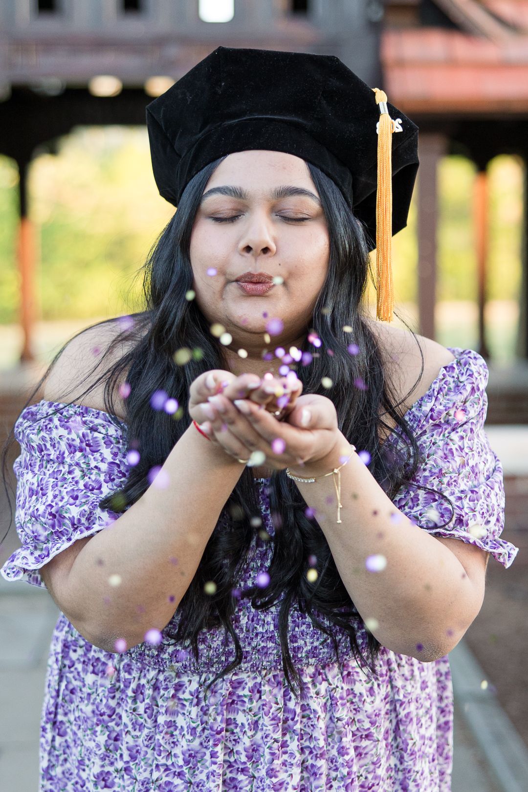 Graduate in purple floral dress blows glitter while wearing graduation cap with closed eyes and joyful expression.