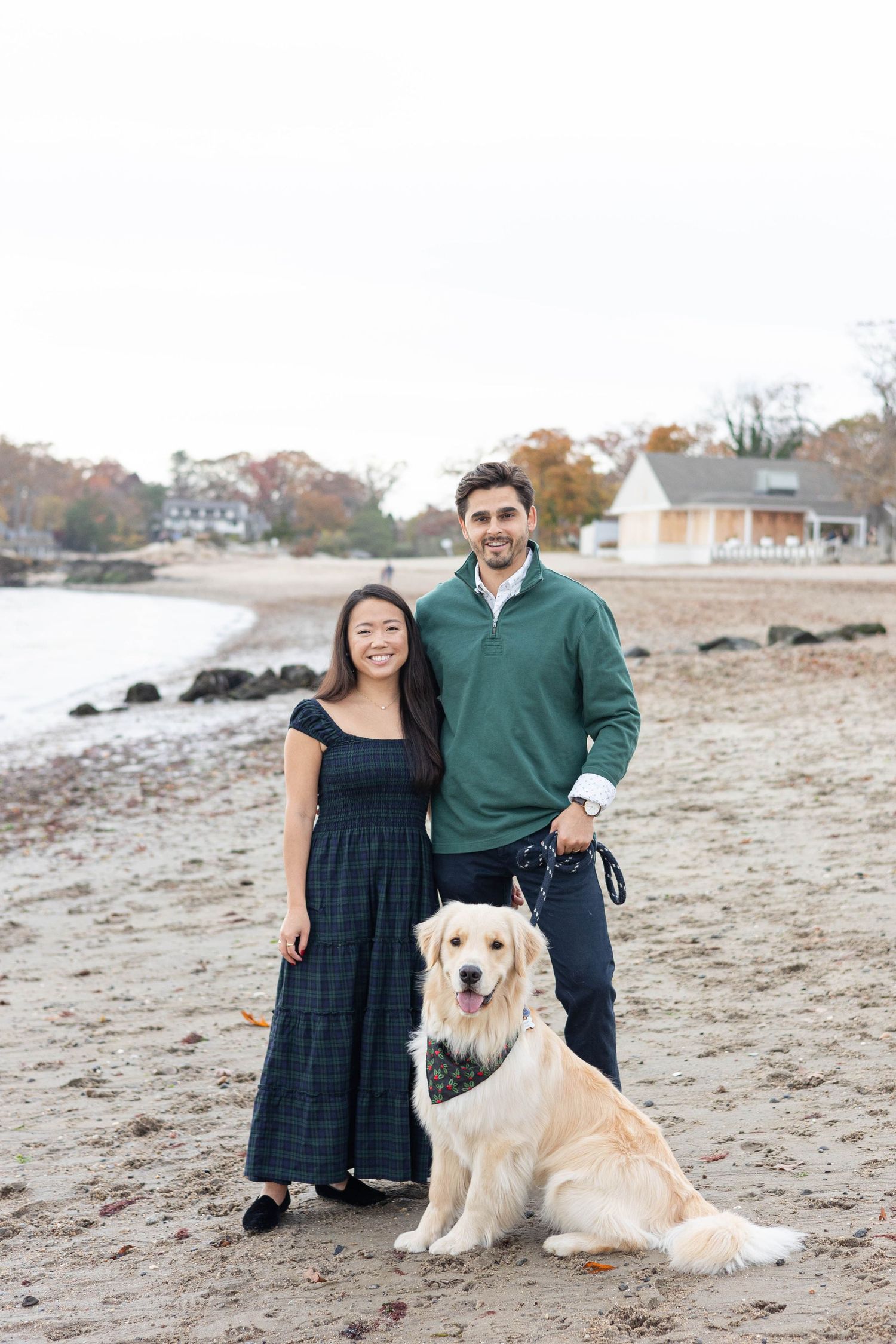 Couple poses with their Golden Retriever on rocky beach with coastal houses in background.