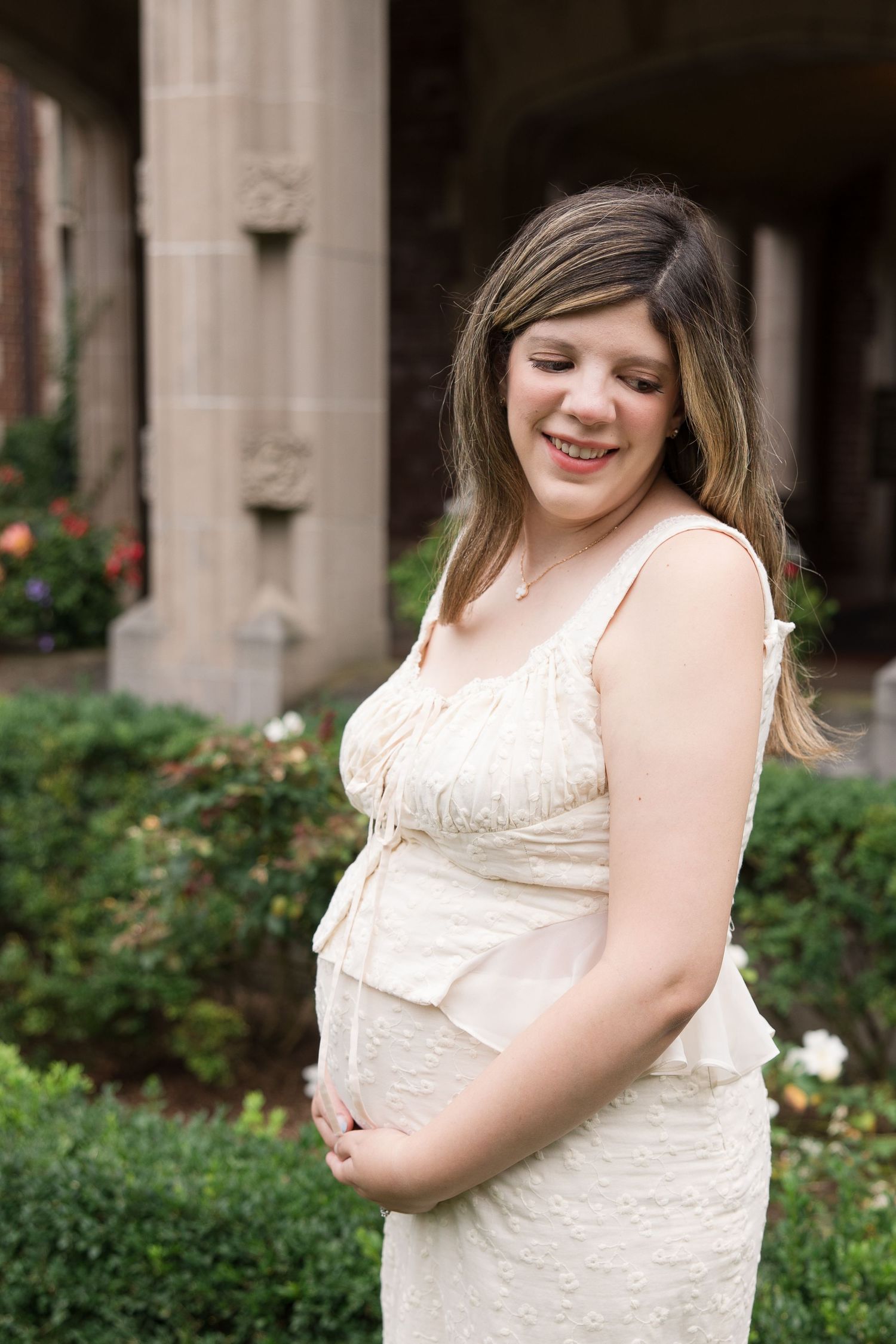 Expecting mother in cream dress smiles while posing near garden archway.