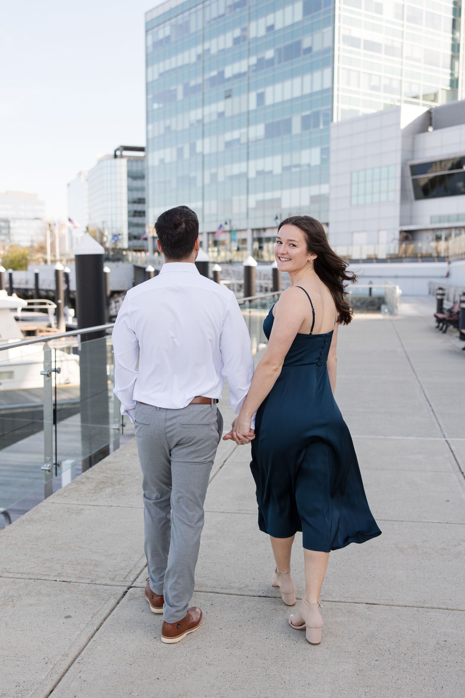A couple walking along a waterfront path with glass buildings in the background.