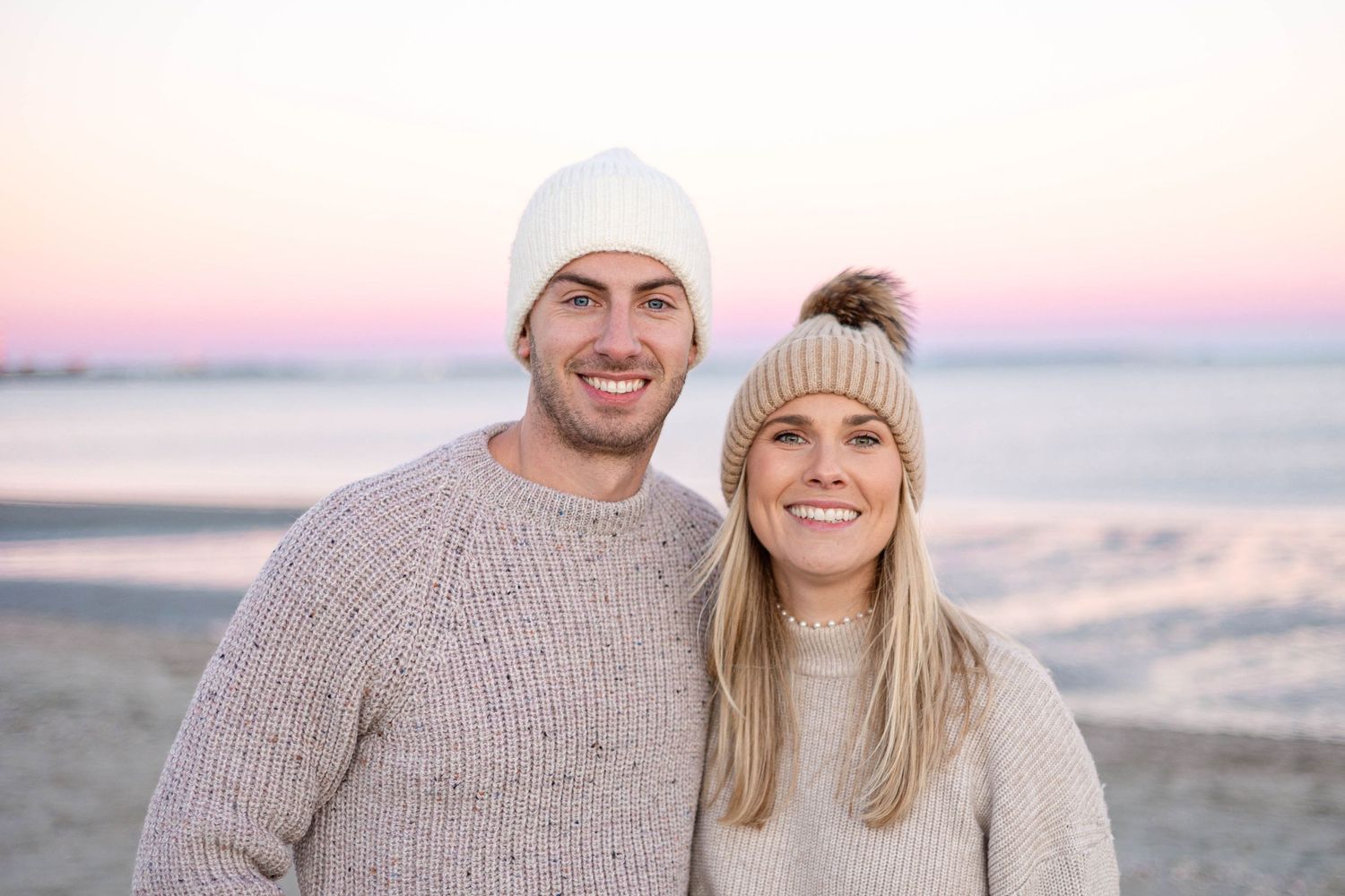 Couple in matching sweaters smile during sunset beach photoshoot.