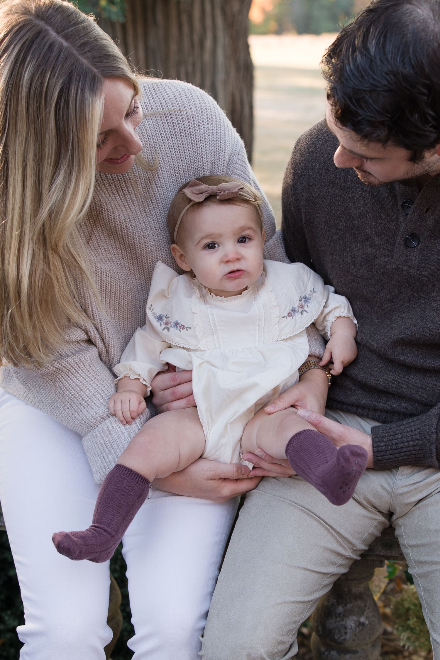 Intimate family moment outdoors with baby wearing white dress and purple socks.