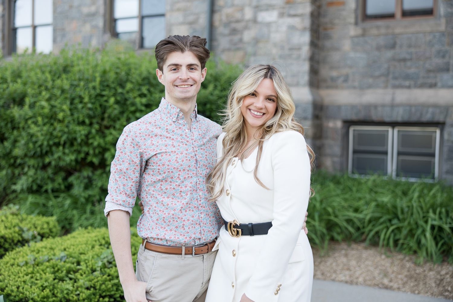 A couple poses outdoors in front of stone architecture and greenery wearing coordinated spring outfits.