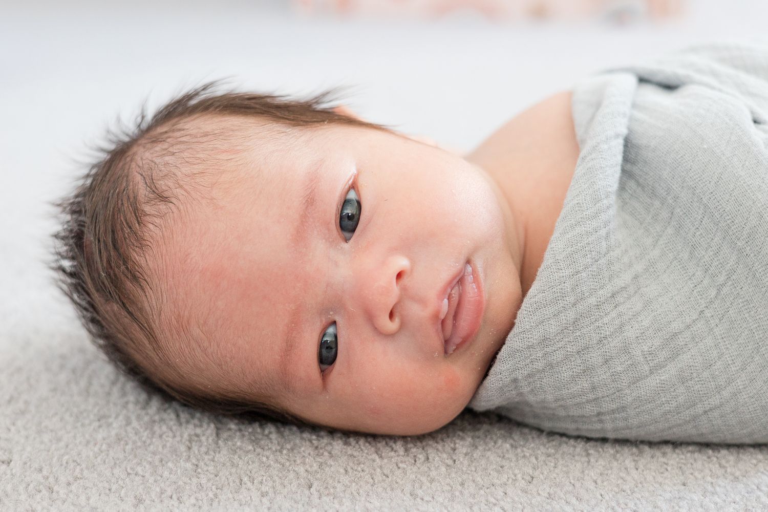 A newborn baby wrapped in gray swaddle smiles while lying on a light gray surface.