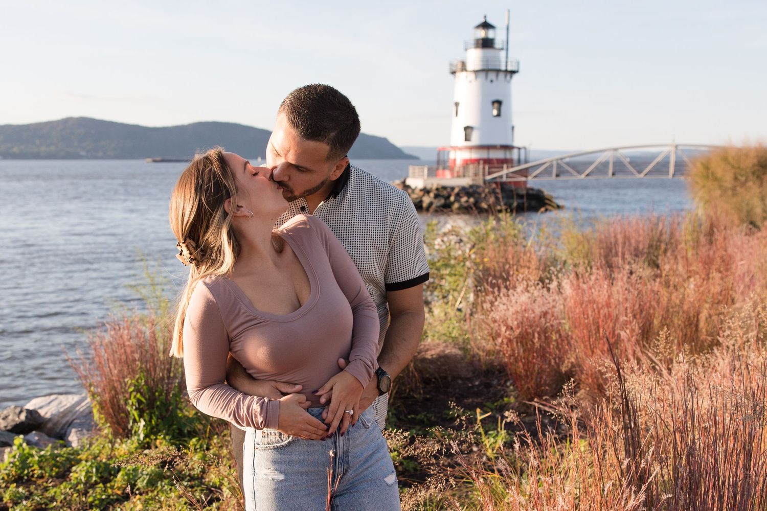 Couple sharing tender moment near lighthouse with mountains and water in background.