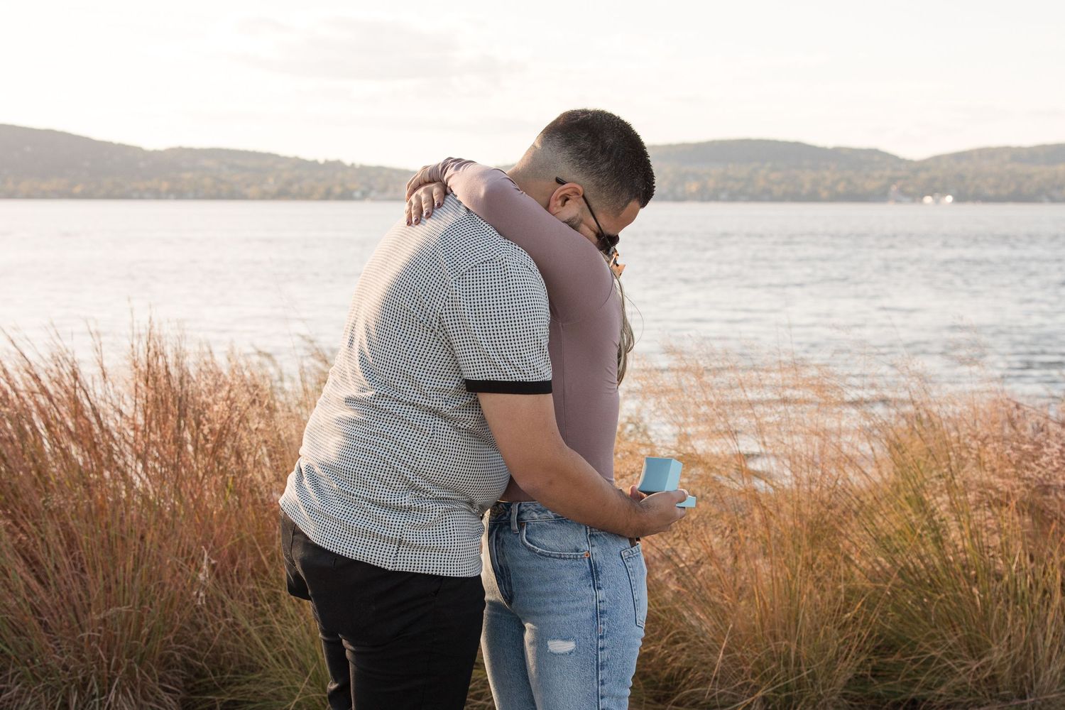 Couple embracing after proposal by lighthouse with water and mountains in background.