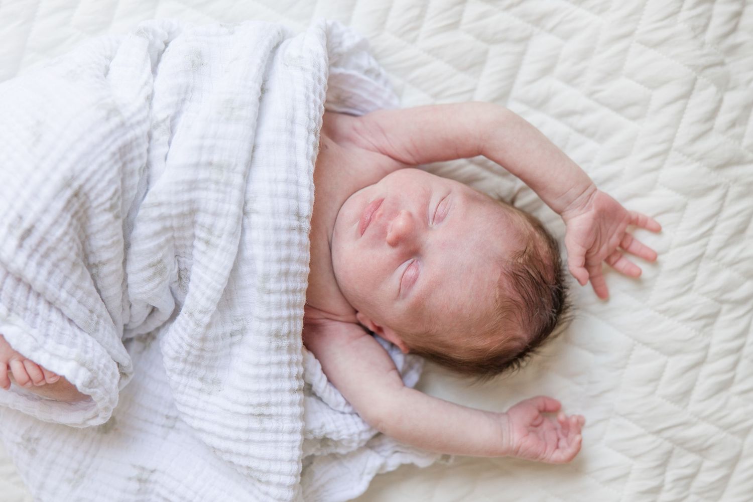 A sleeping newborn baby swaddled in white cloth rests peacefully on a textured white blanket.