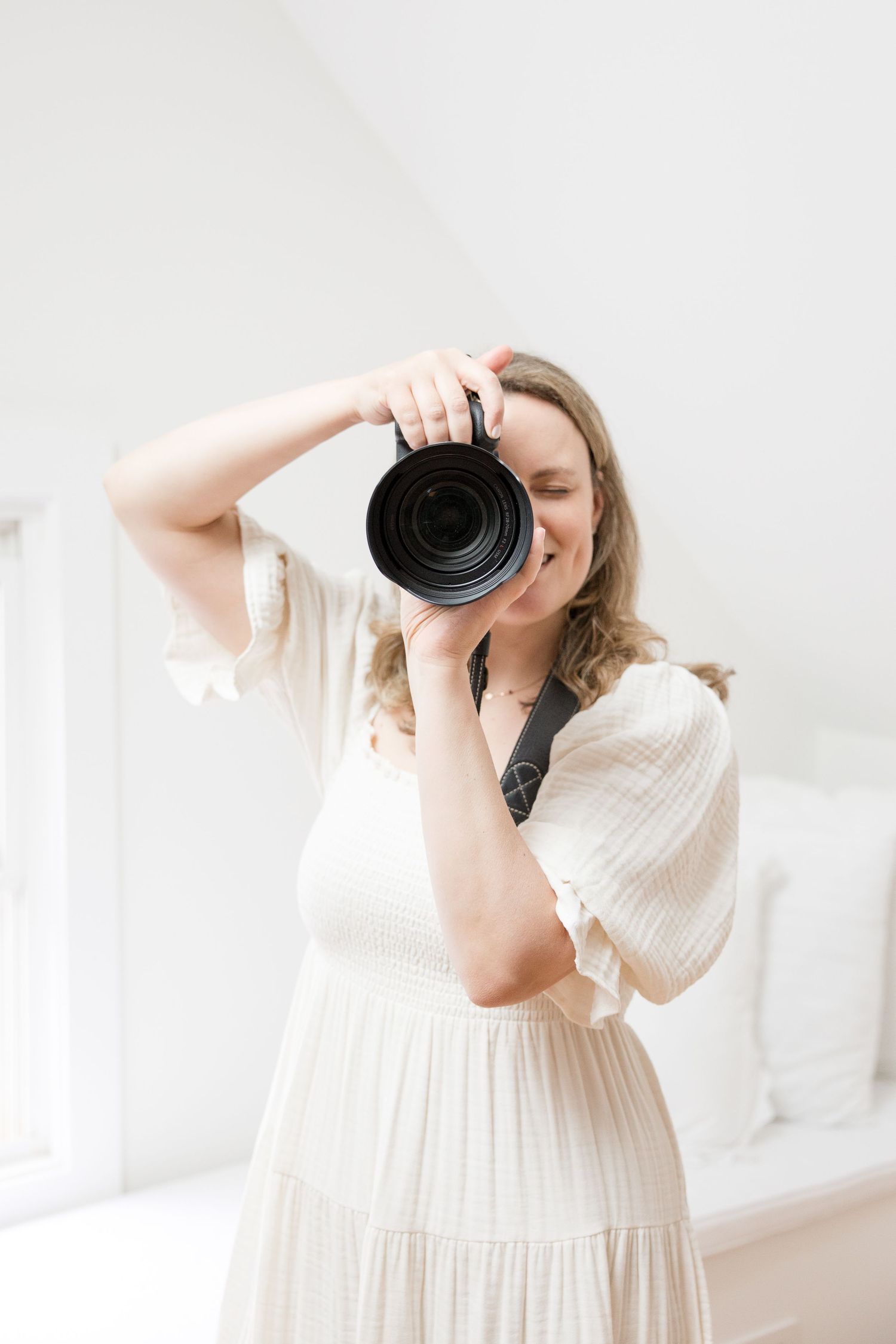 Photographer in flowing white dress holds professional camera in bright minimal interior space.
