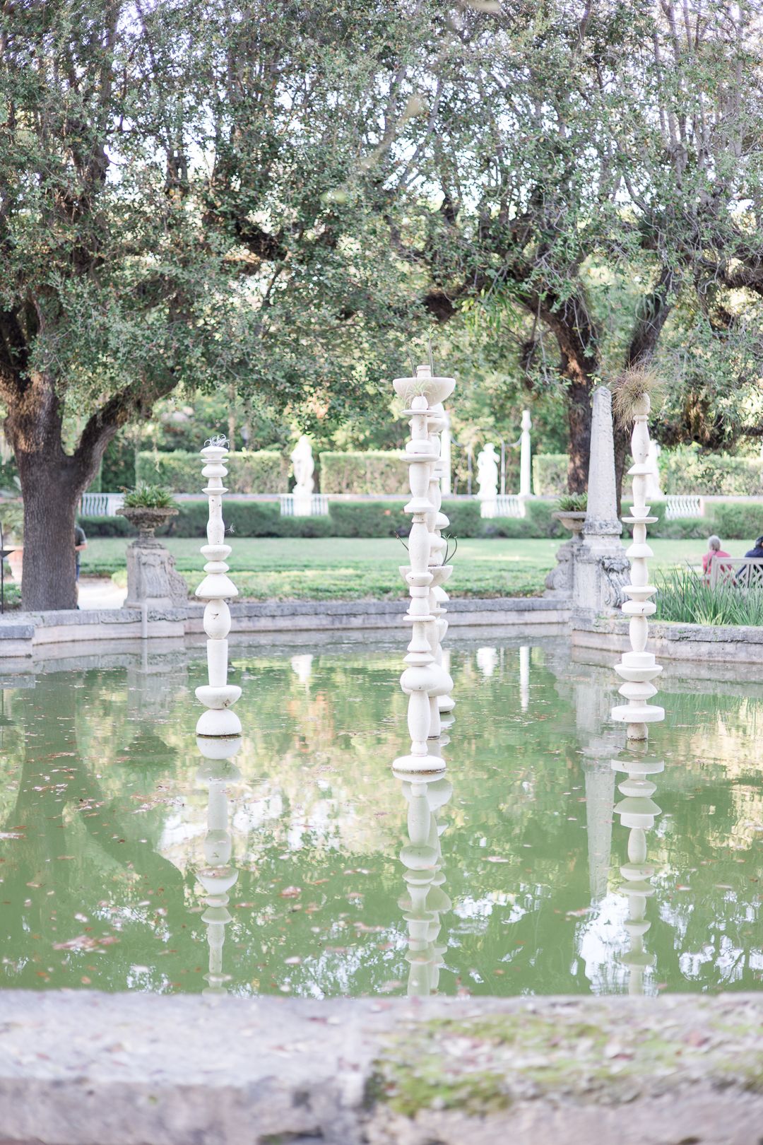 Ornate stone pillars stand in a reflective pond surrounded by trees in a serene garden setting.