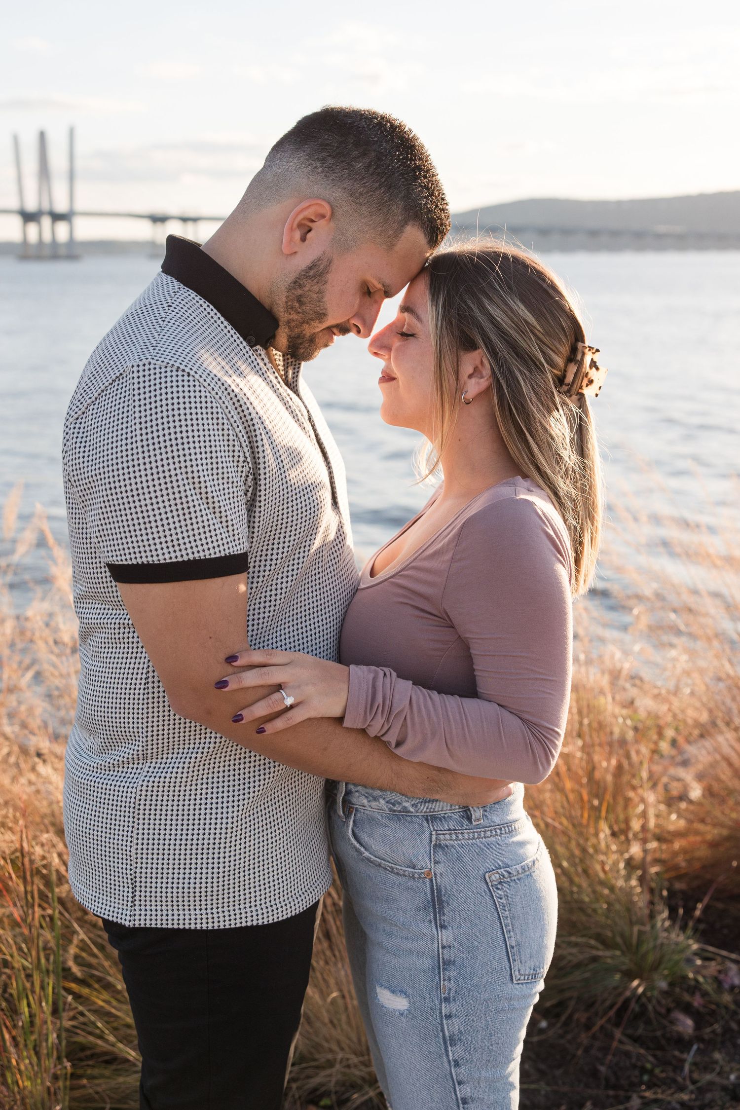 Intimate portrait of couple embracing with bridge visible in background.