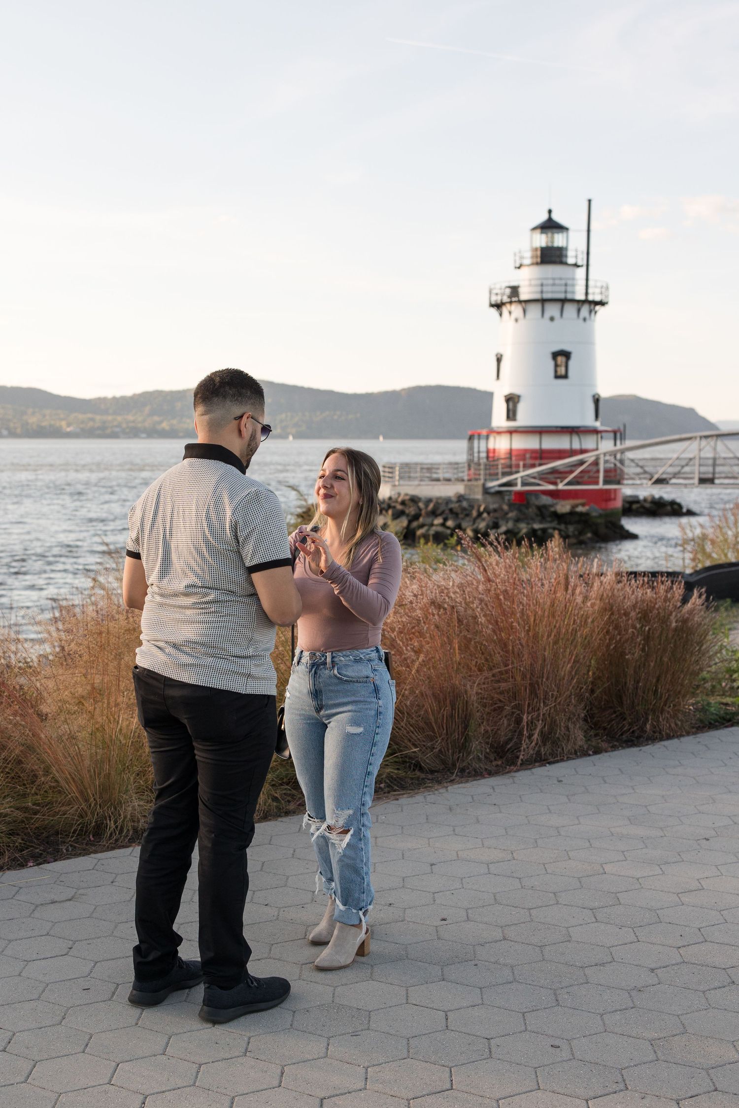 A couple shares a romantic moment in front of a lighthouse on the water during sunset.
