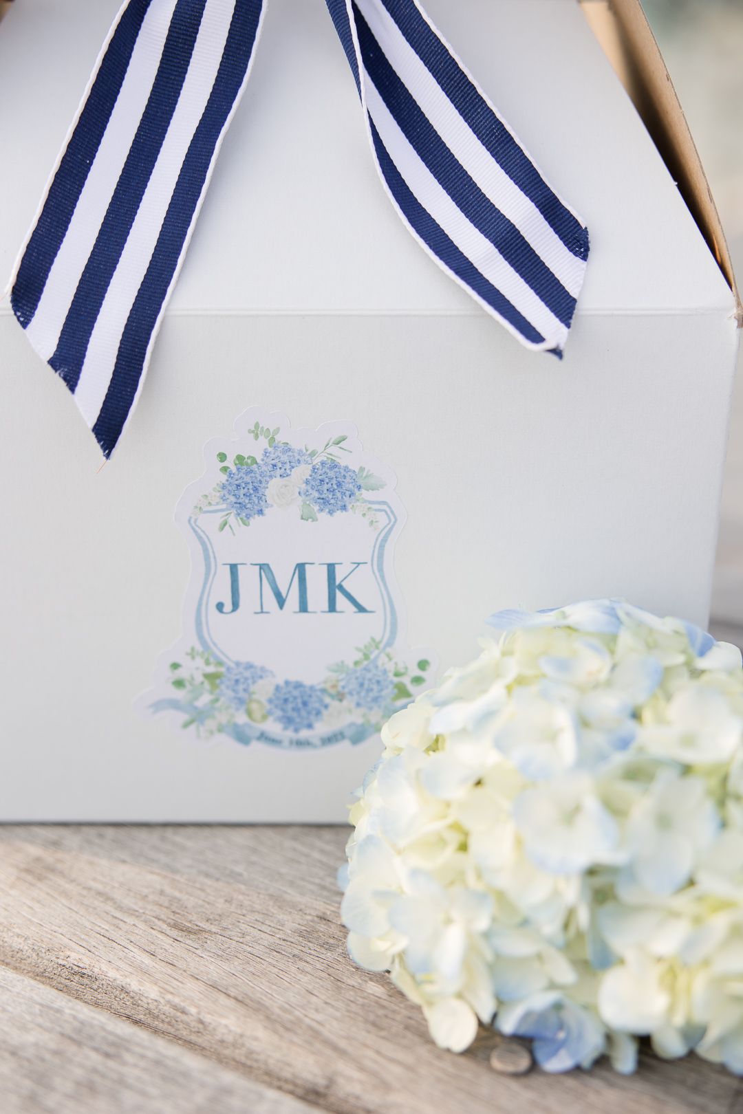 White gift box with navy striped ribbon and JMK monogram next to hydrangea flower.