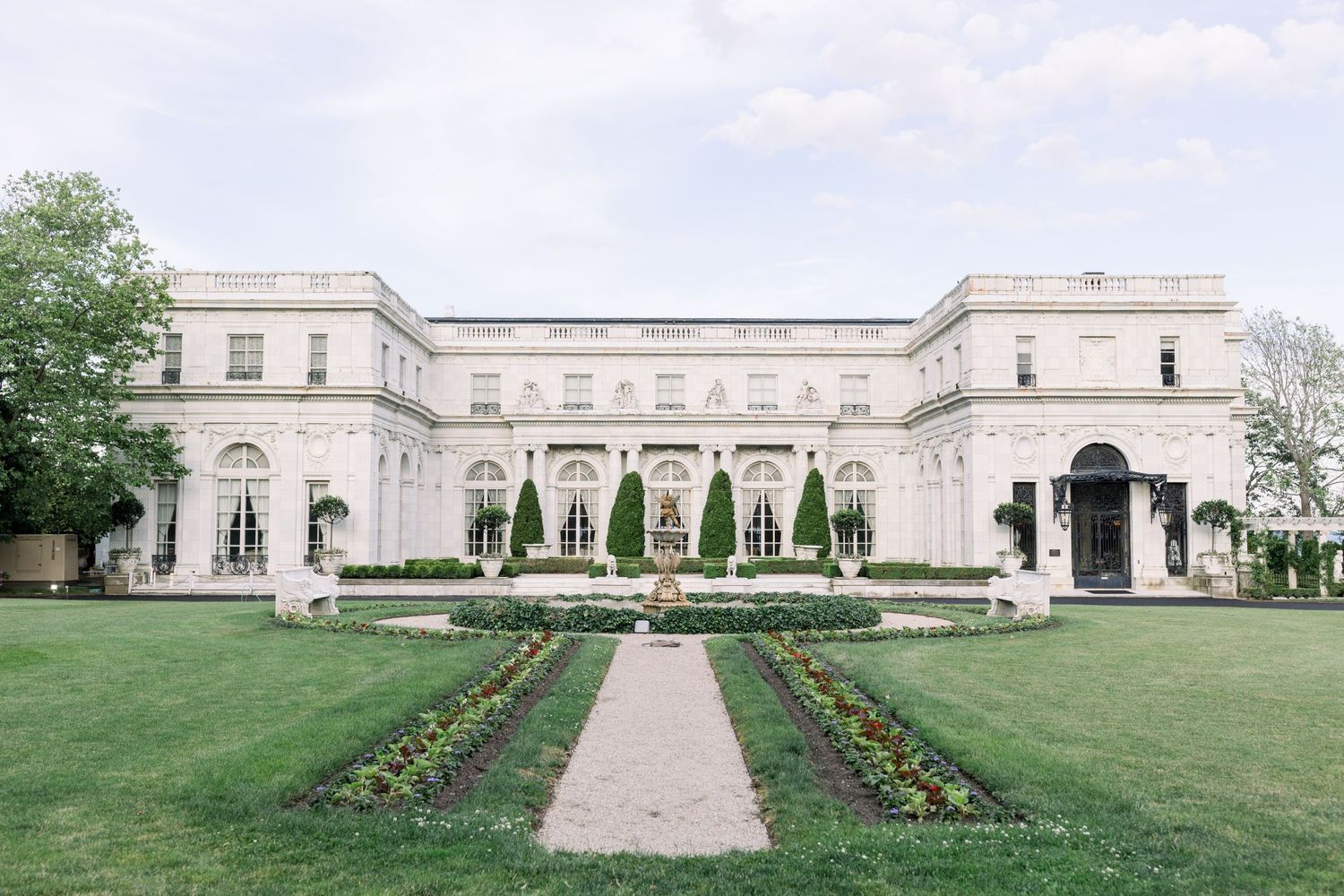 Elegant white mansion with manicured gardens and floral lined walkway leading to grand entrance.