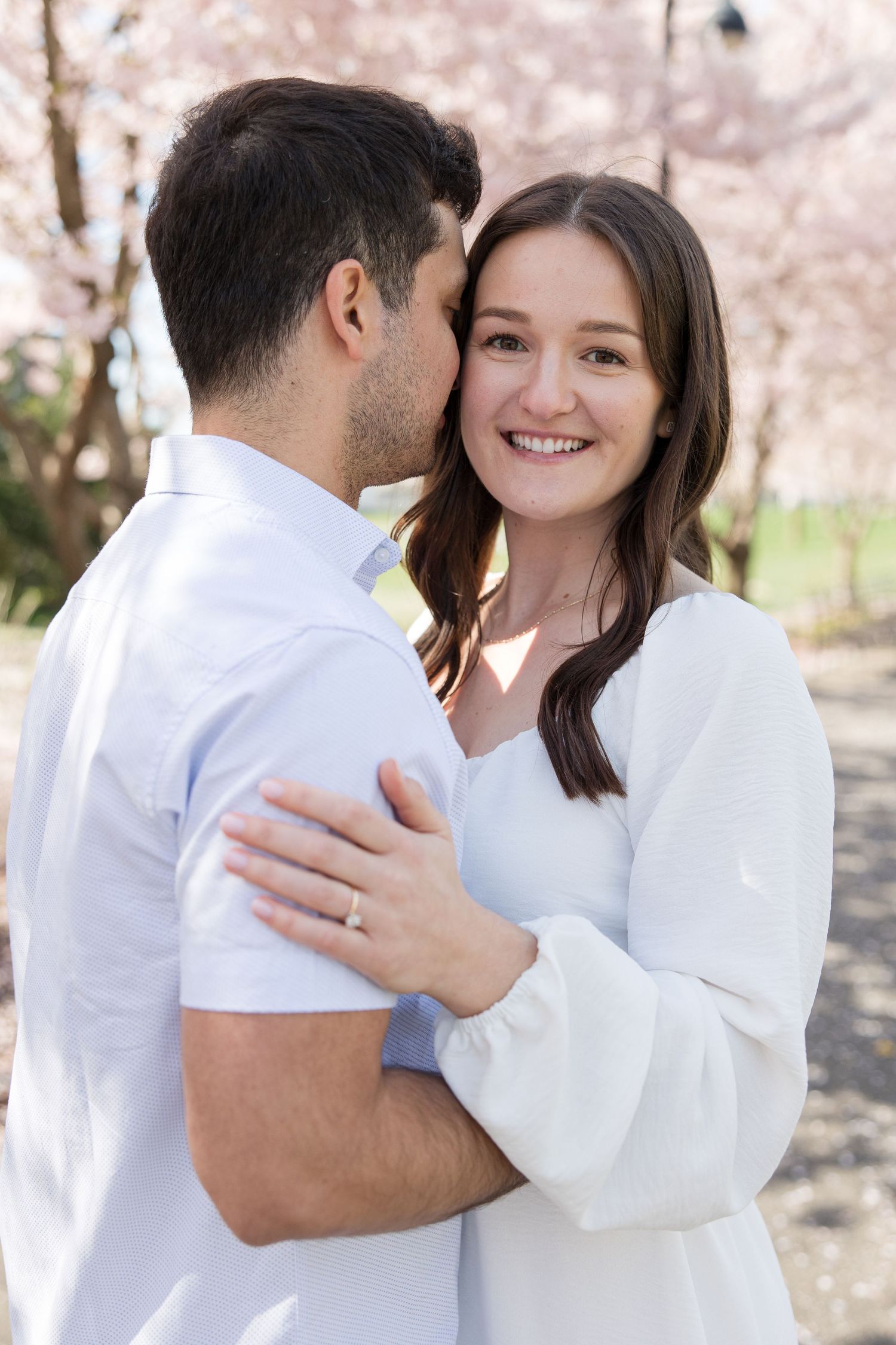 Happy engaged couple in white embracing under cherry blossom trees.