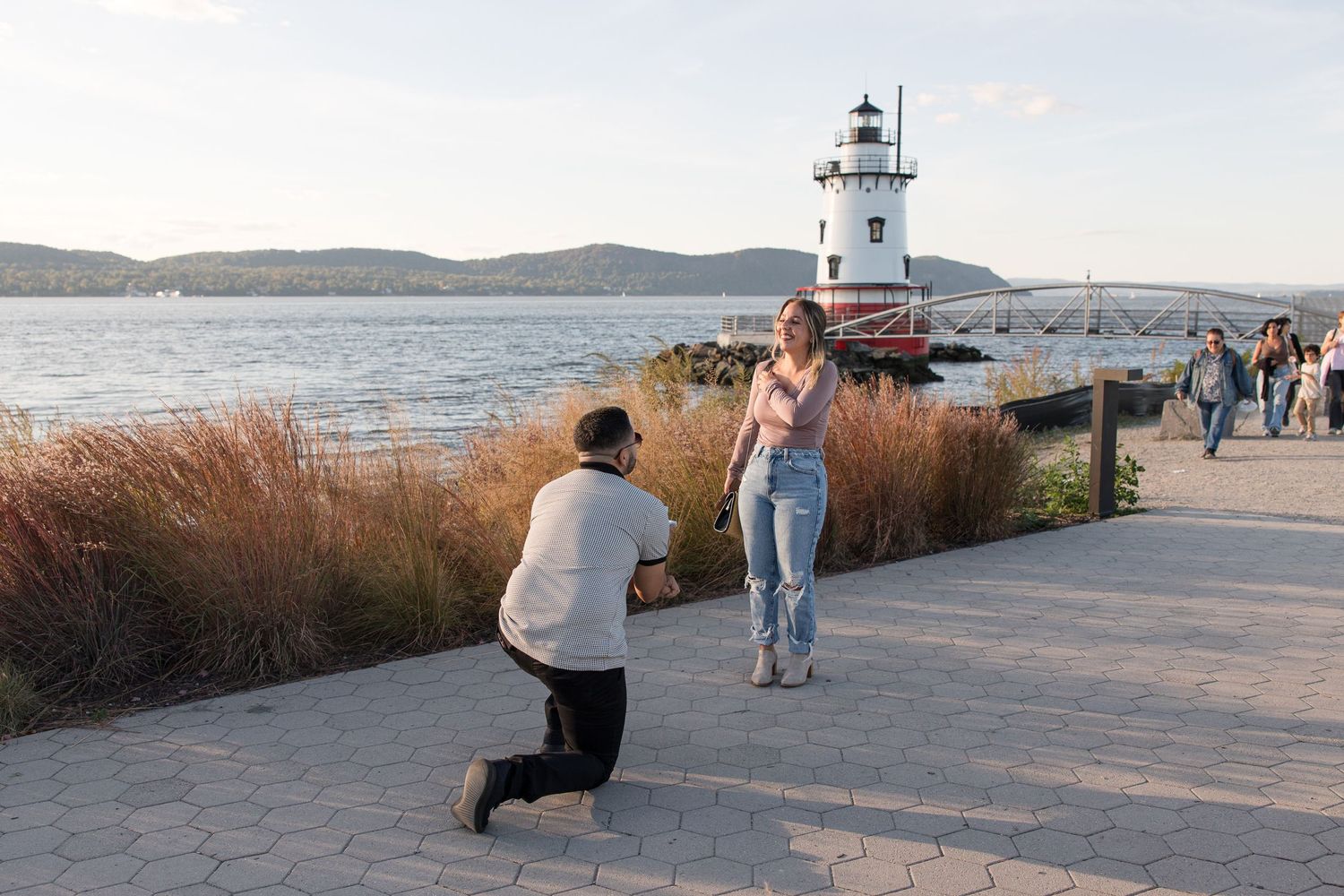 Surprise proposal at waterfront lighthouse path during sunset.